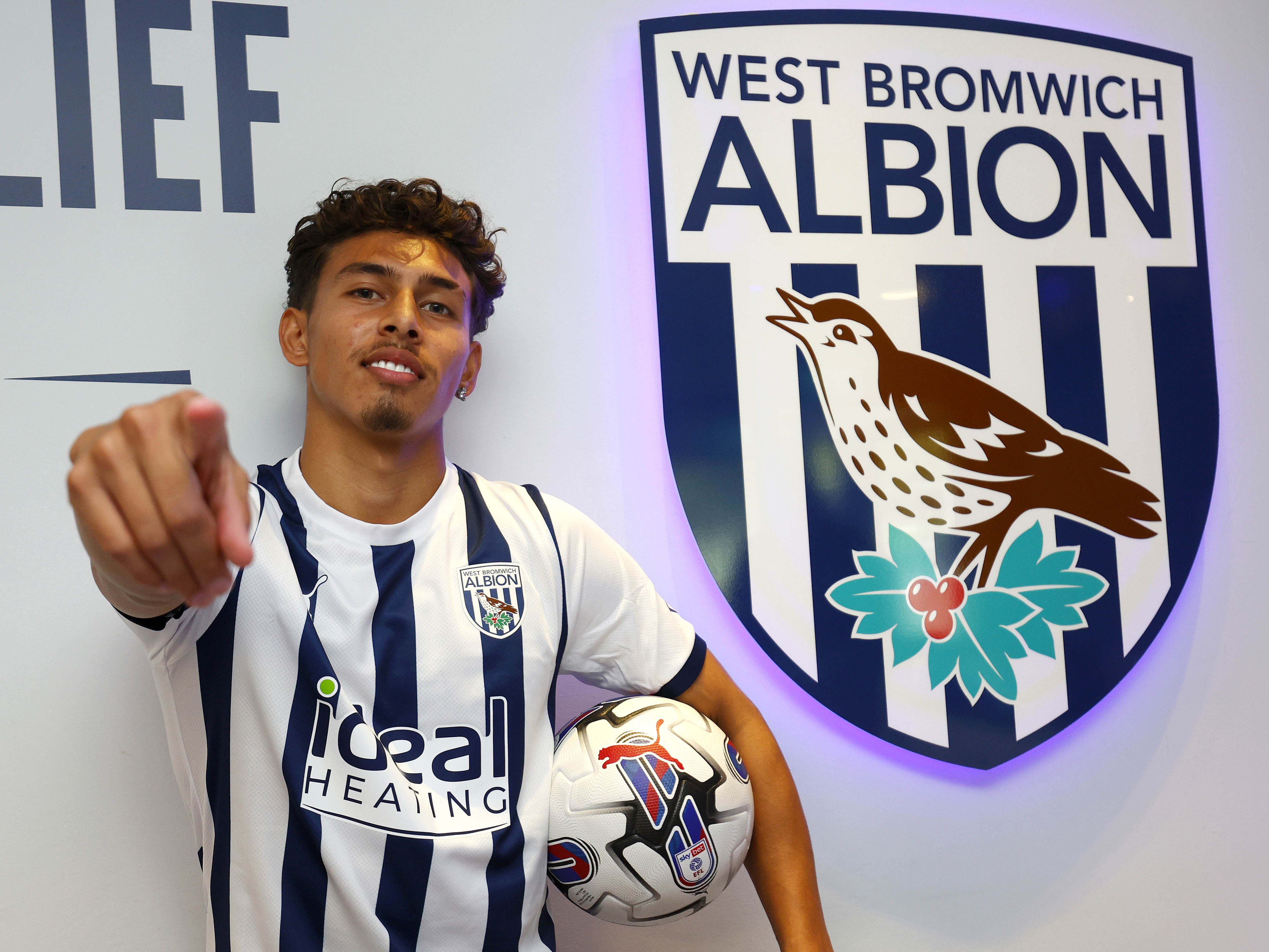 Jeremy Sarmiento next to a WBA badge pointing at the camera holding a football