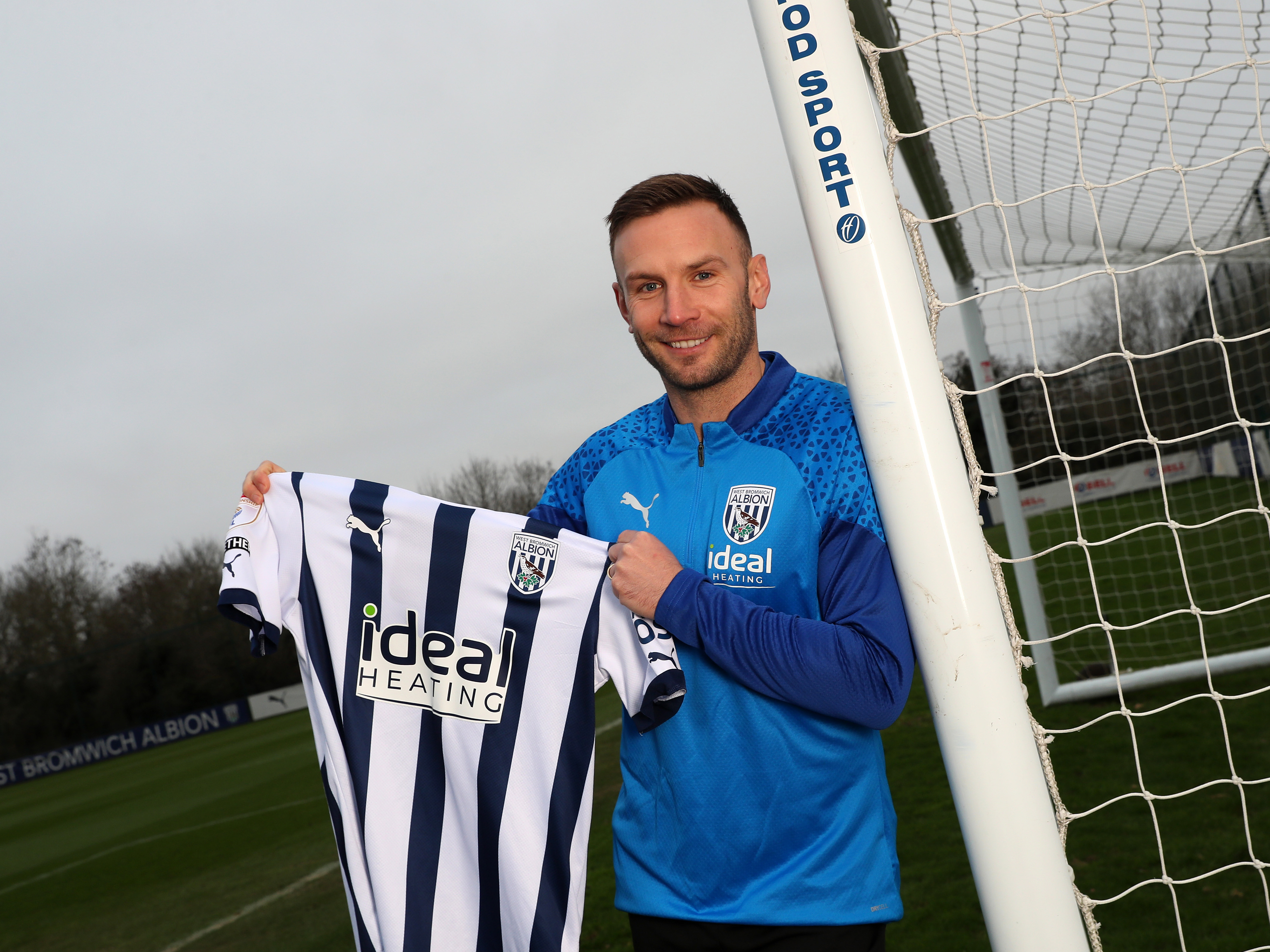 Andi Weimann smiling at the camera, leaning up against a goal post and holding a WBA home shirt up
