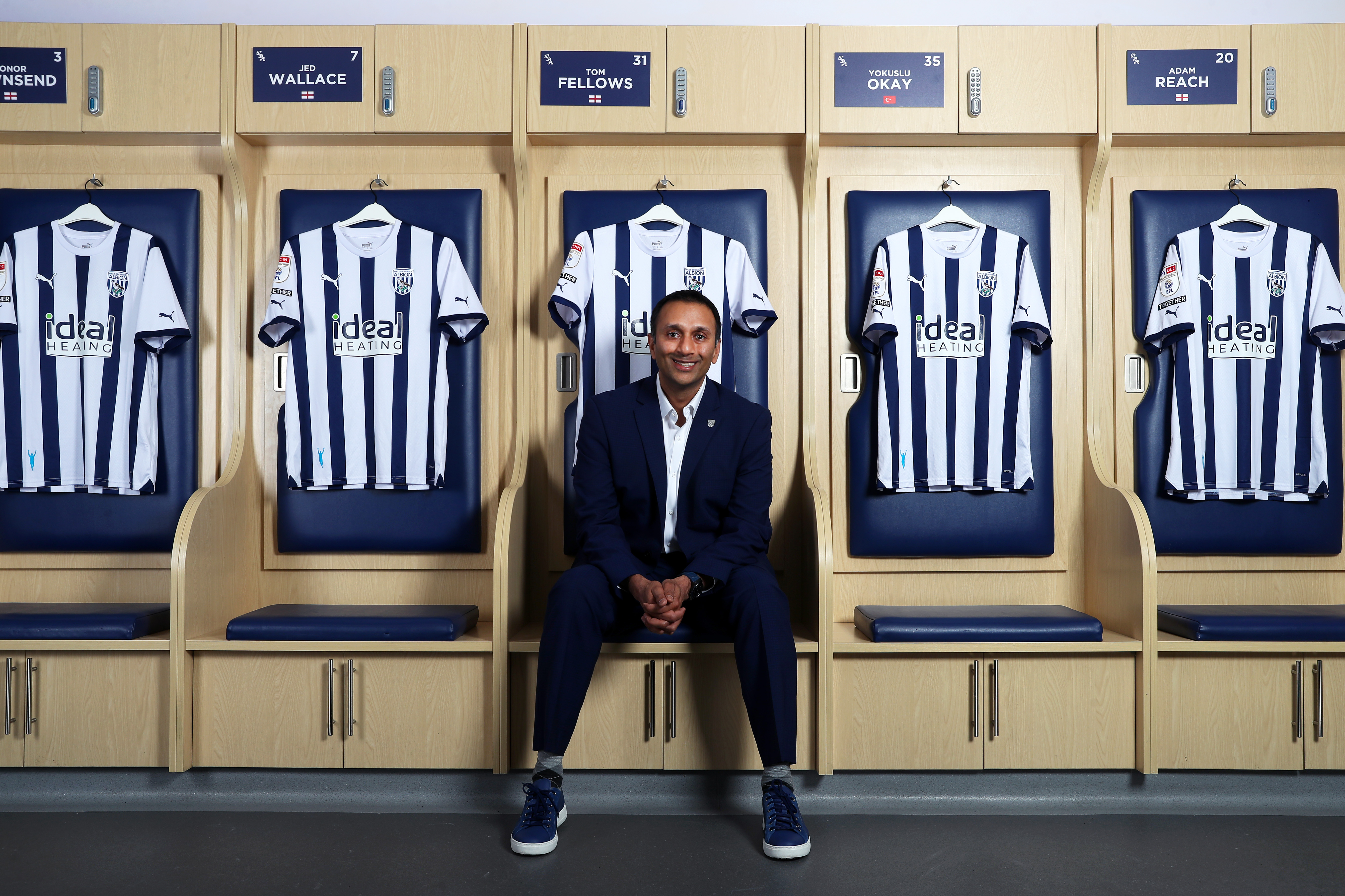 Shilen Patel sat in the home dressing room smiling at the camera with several home shirts hanging up behind him
