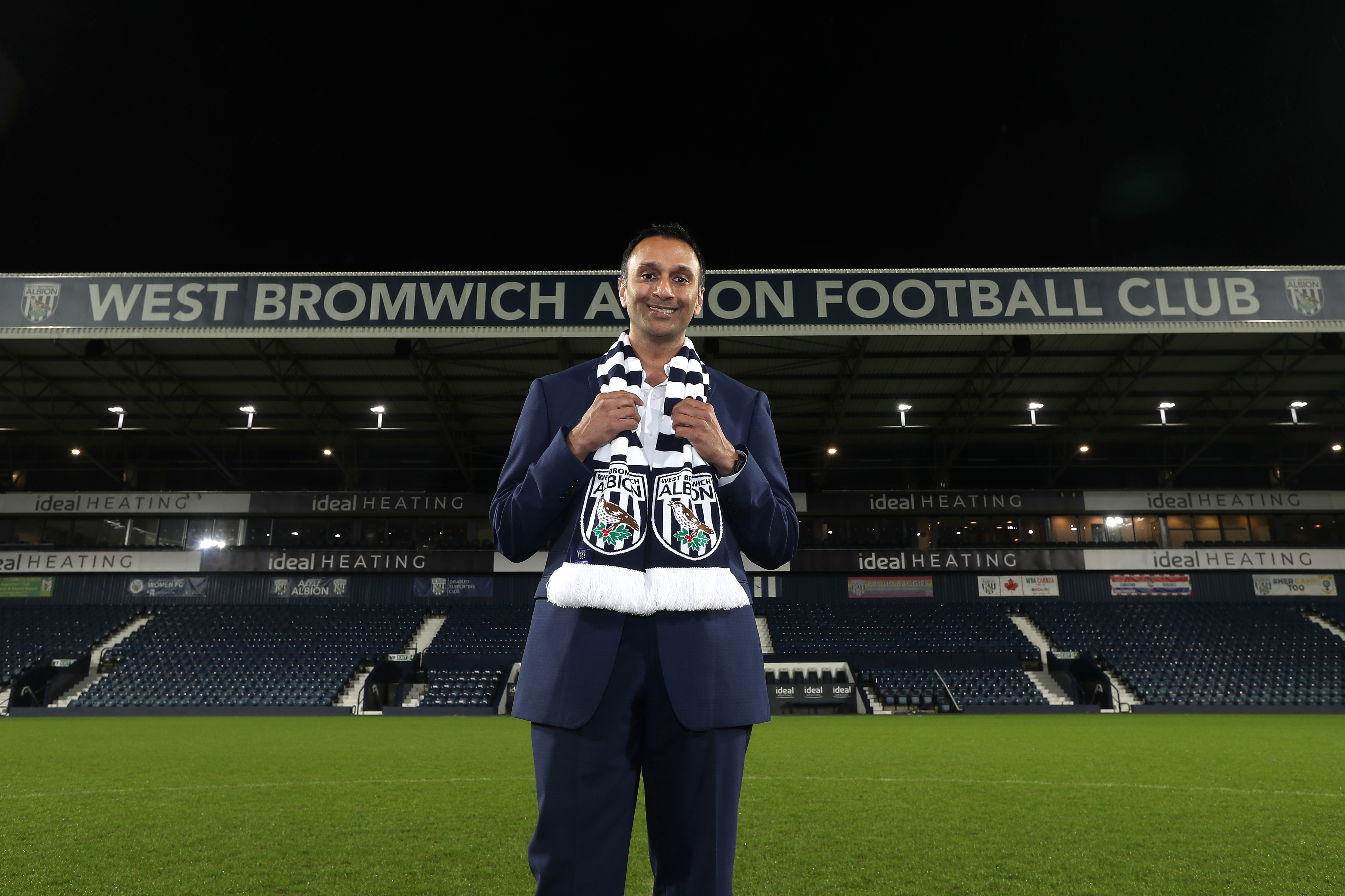 Shilen Patel stood on the pitch at The Hawthorns with the West Stand behind him and an Albion scarf wrapped around his neck