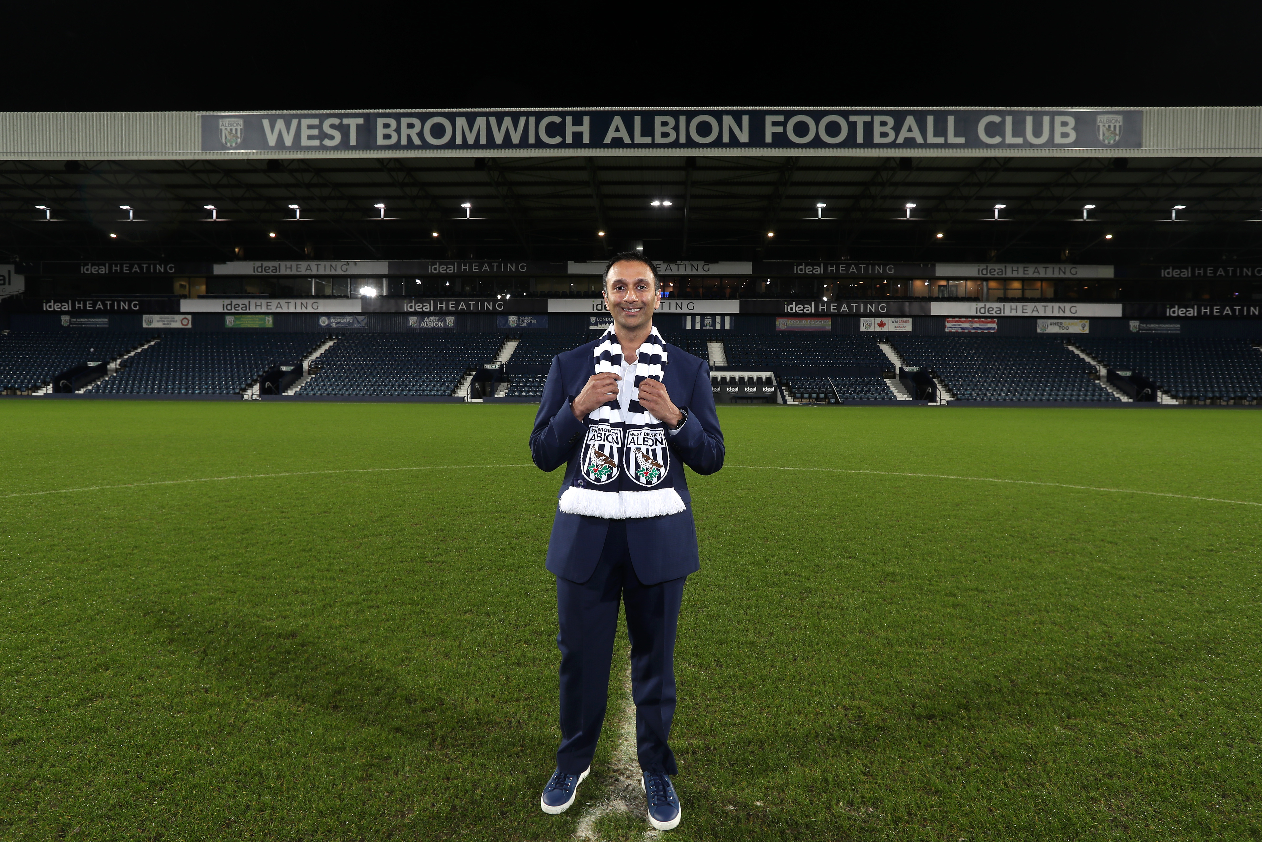 Shilen Patel stood on the pitch at The Hawthorns smiling at camera with the West Stand in the background and an Albion scarf wrapped round his neck