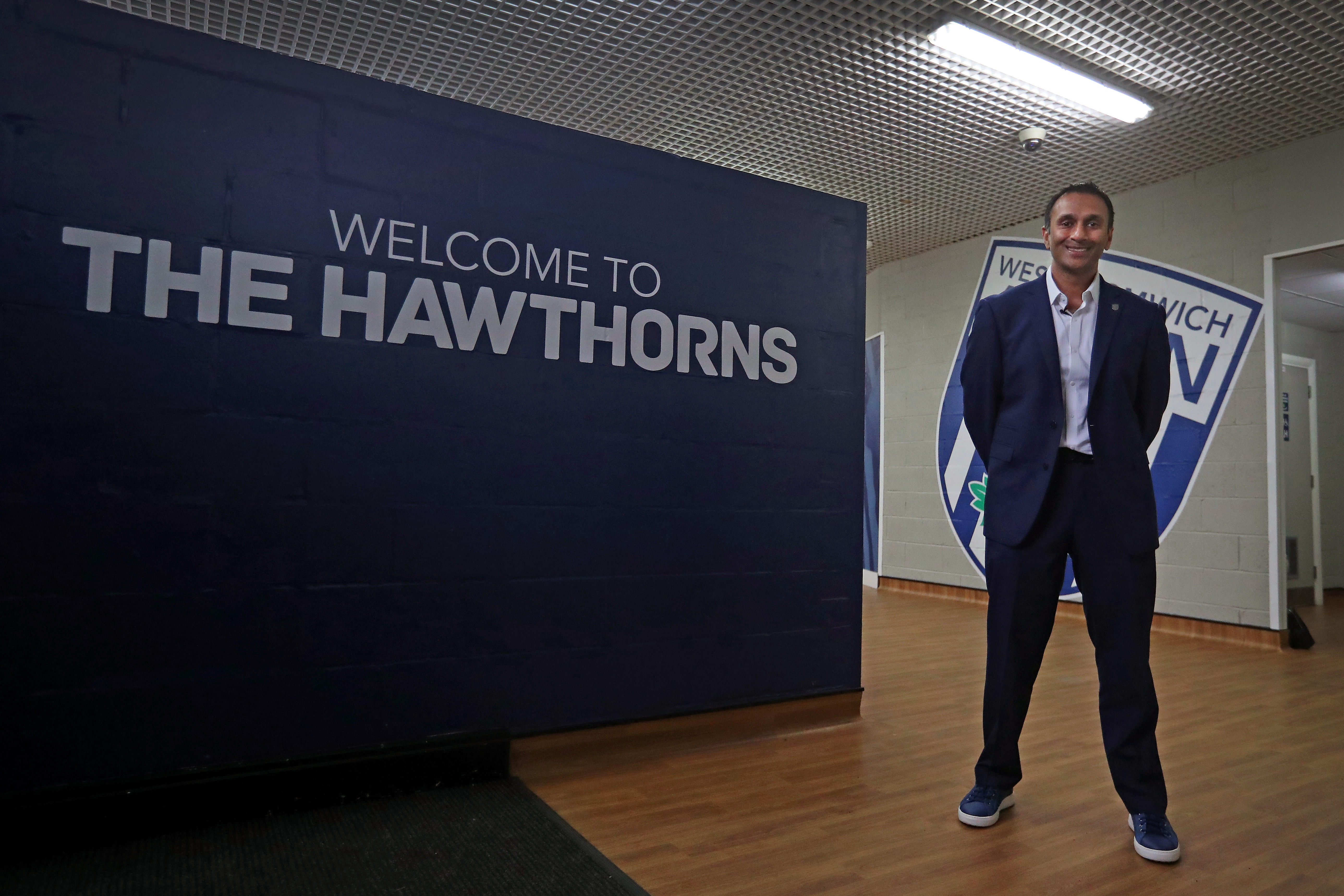 Shilen Patel stood in a suit next to a wall with "Welcome to The Hawthorns" written on it