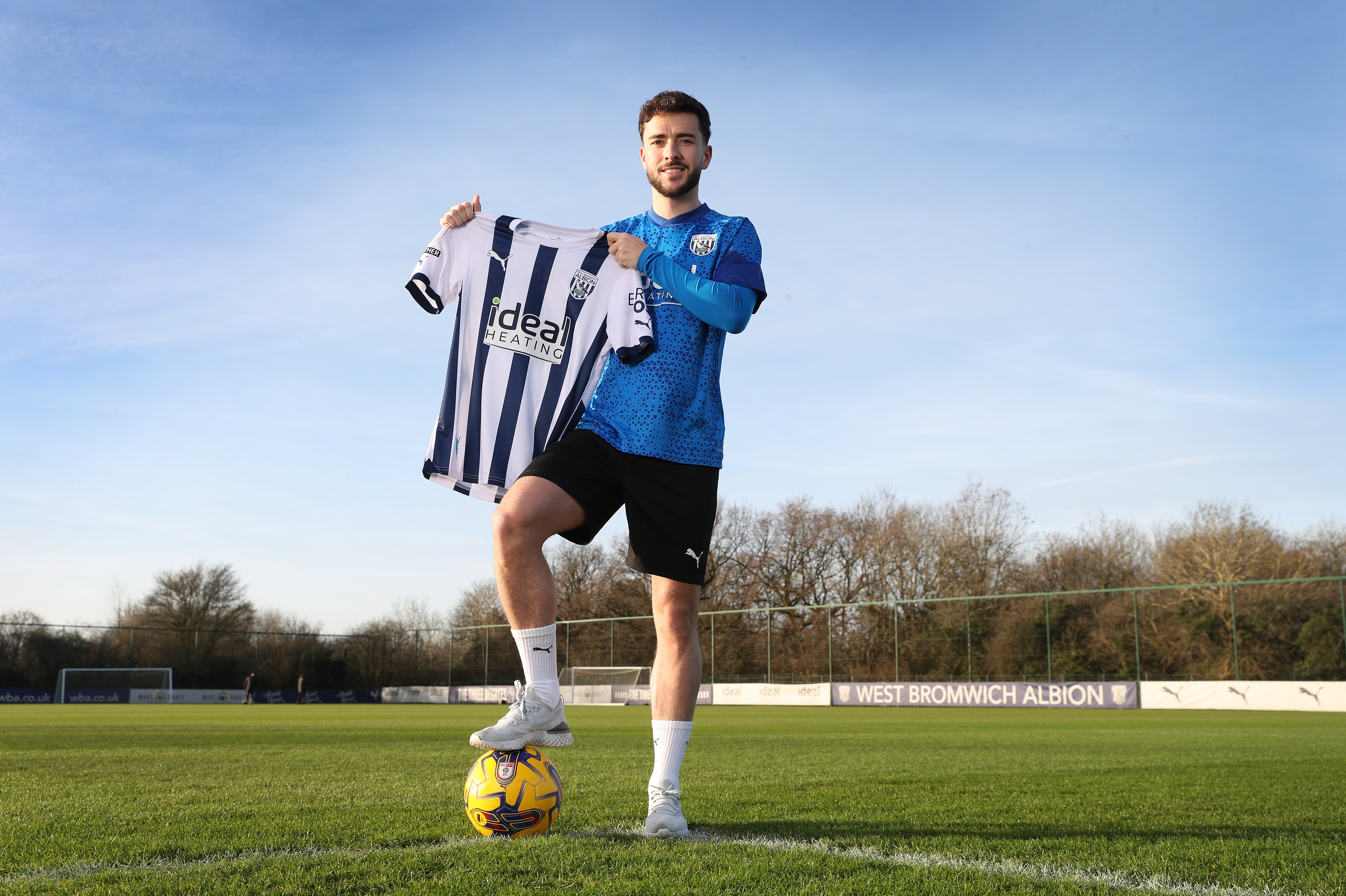 Mikey Johnston posing for a photo with one foot on a yellow football while holding a home Albion shirt