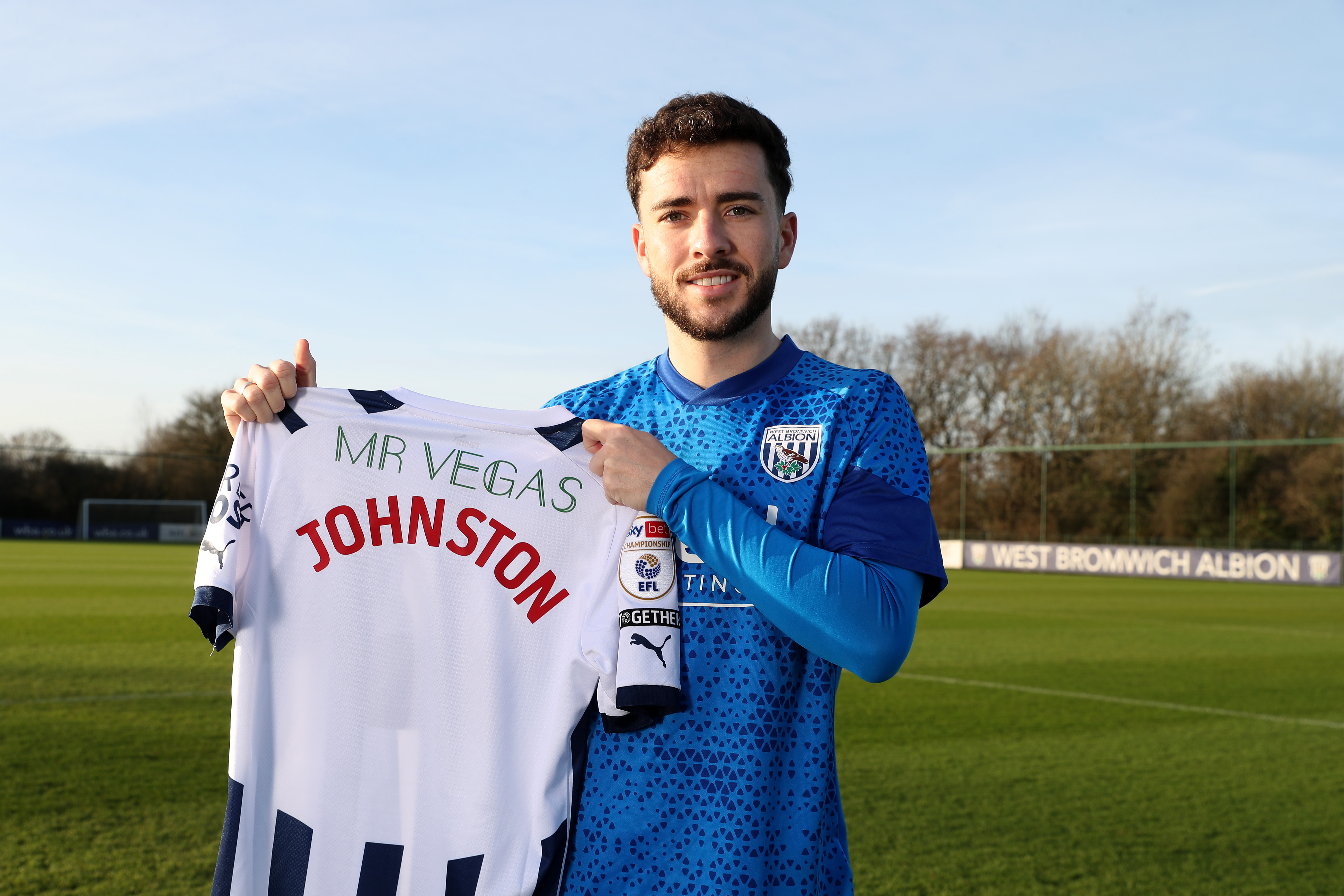 Mikey Johnston posing for a photo outside, smiling and holding up the back of a home shirt with his surname on