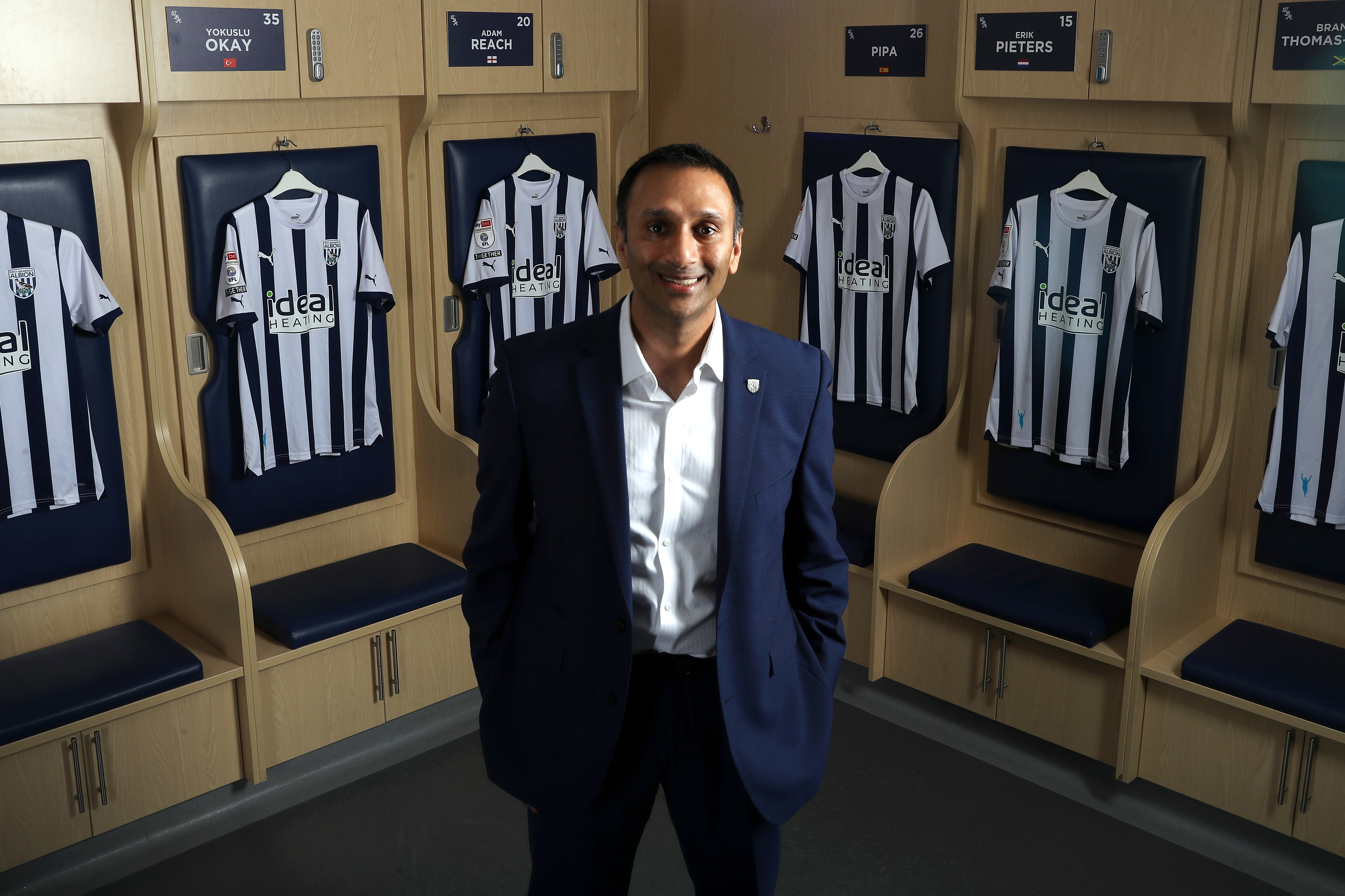 Shilen Patel standing in a suit in the home dressing room surrounded by Albion shirts