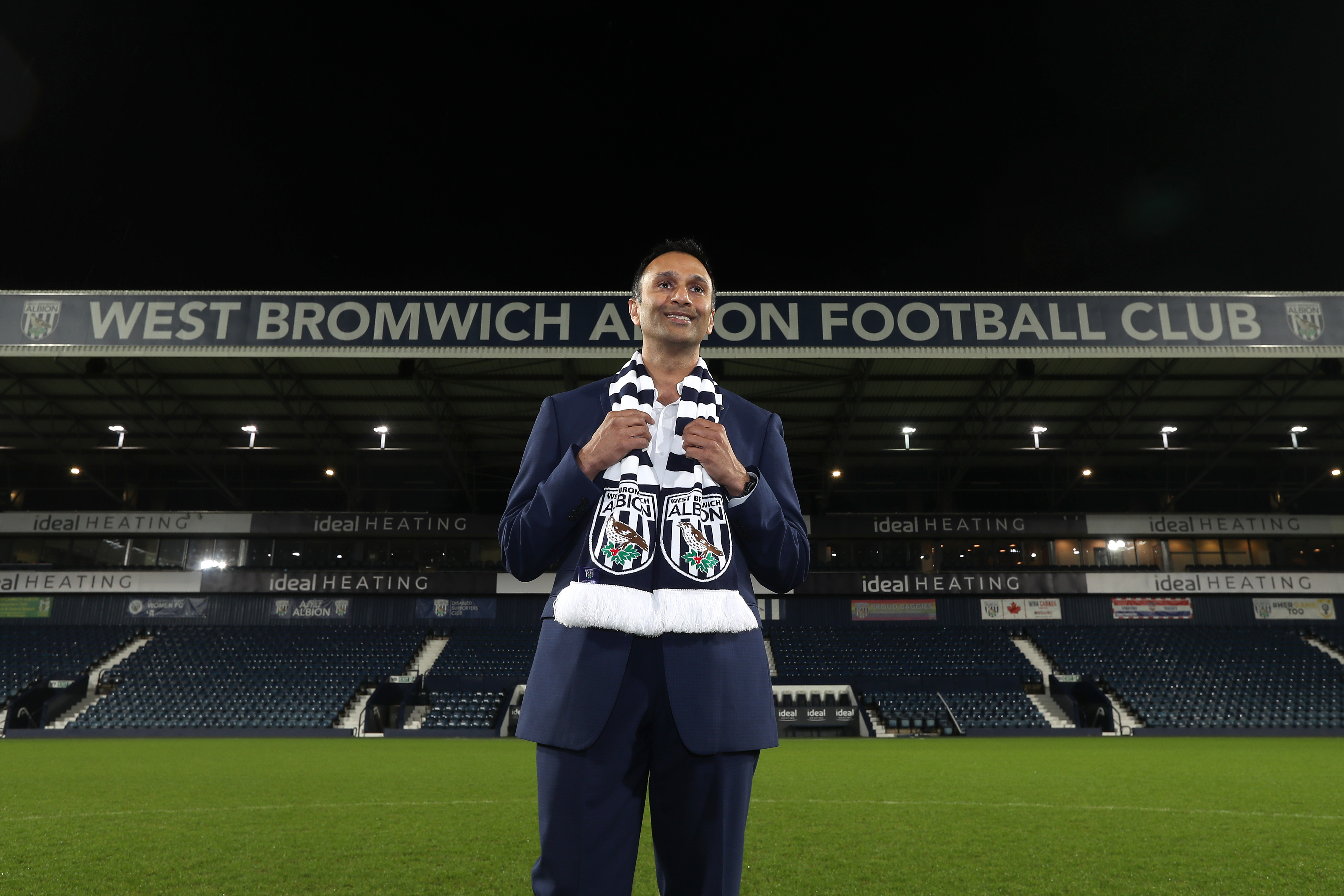 Shilen Patel standing on the pitch at The Hawthorns with the Halfords Lane stand in the background with an Albion scarf around his neck