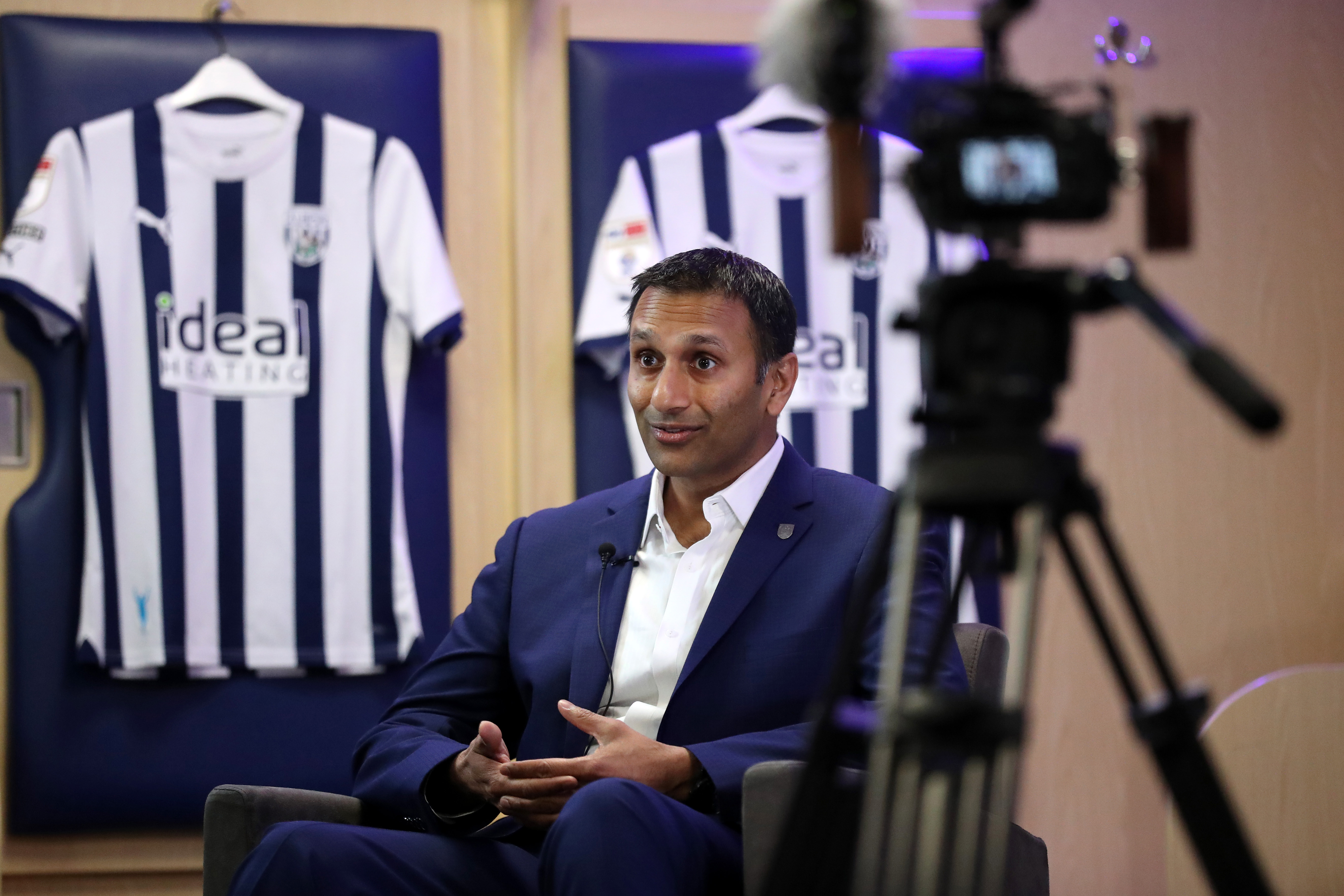 Shilen Patel is interviewed by WBA TV in the home dressing room at The Hawthorns with Albion shirts behind him and a camera to his side