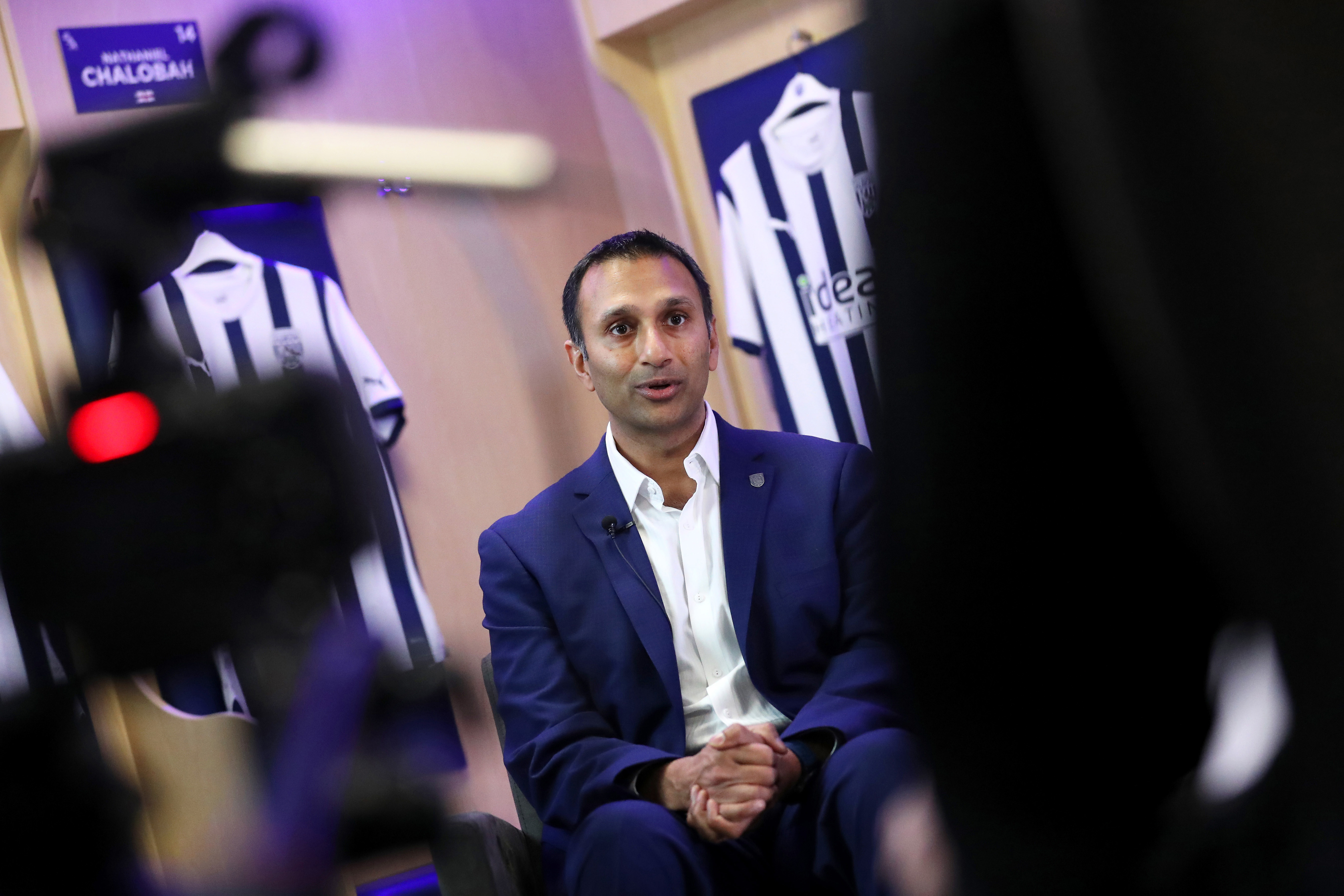 Shilen Patel is interviewed by WBA TV in the home dressing room at The Hawthorns with Albion shirts behind him and a camera to his side