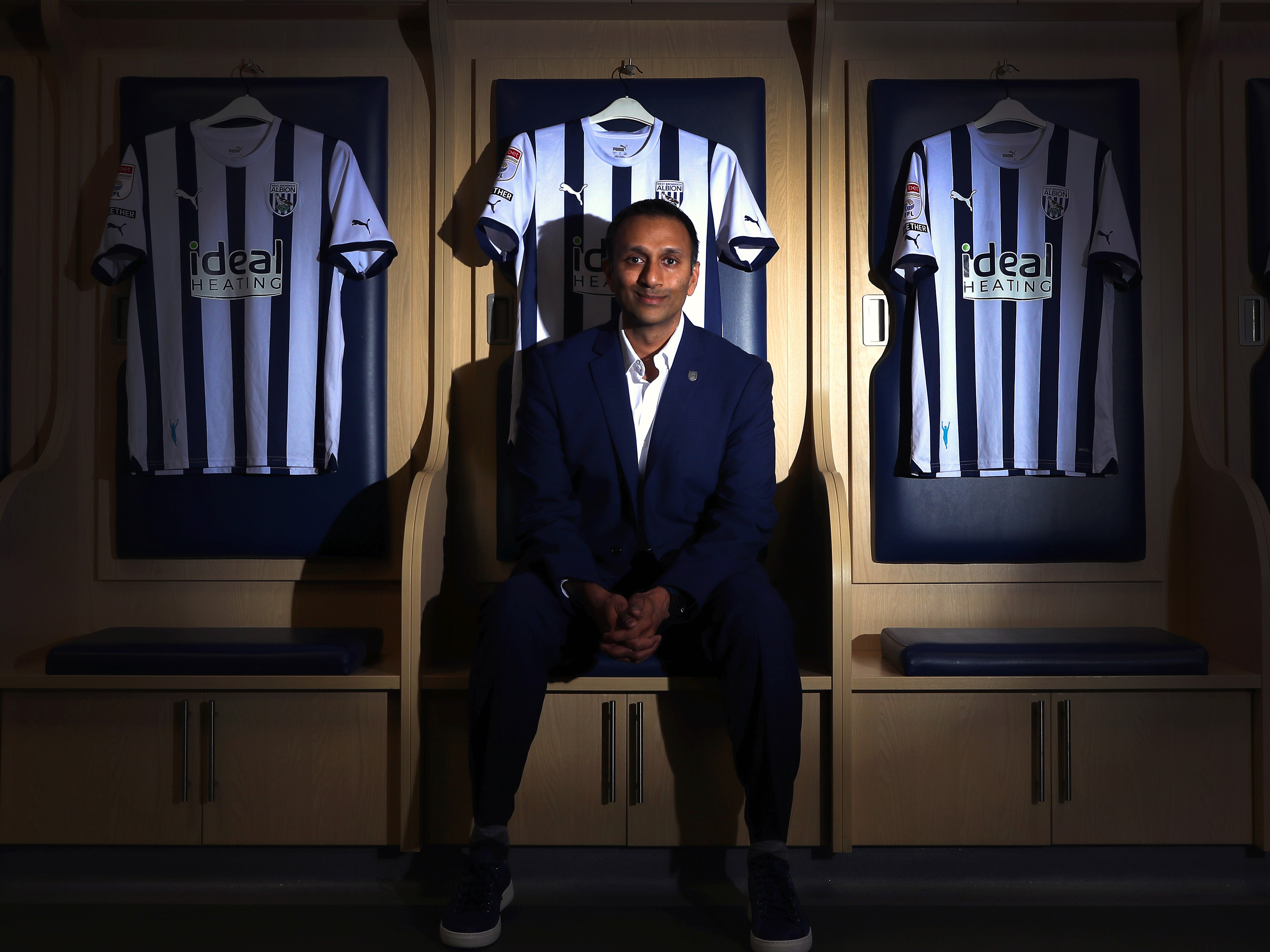 Shilen Patel sitting in the home dressing room looking at the camera surrounded by WBA home shirts