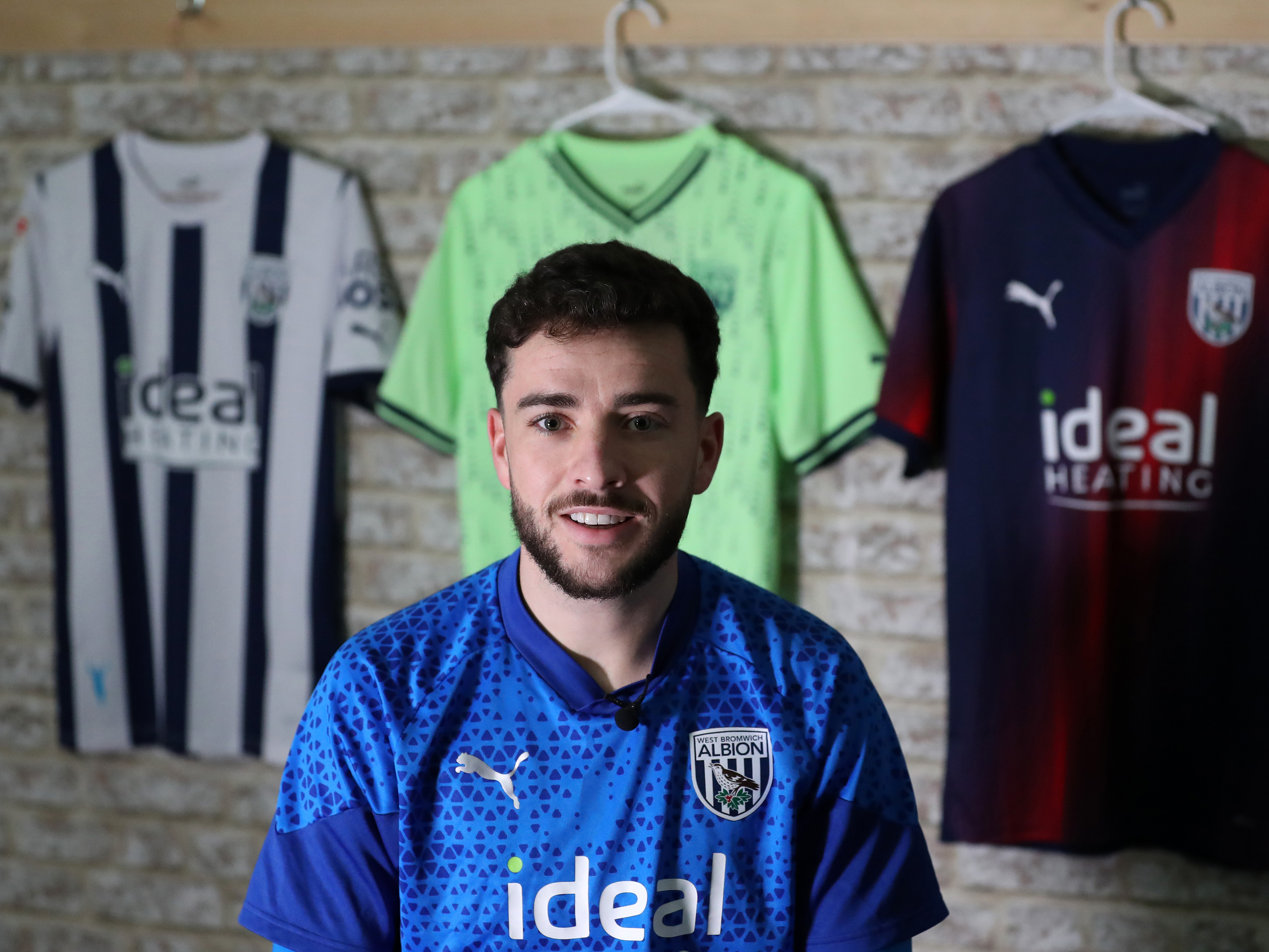 Mikey Johnston smiling at the camera with all three Albion shirts hanging behind him