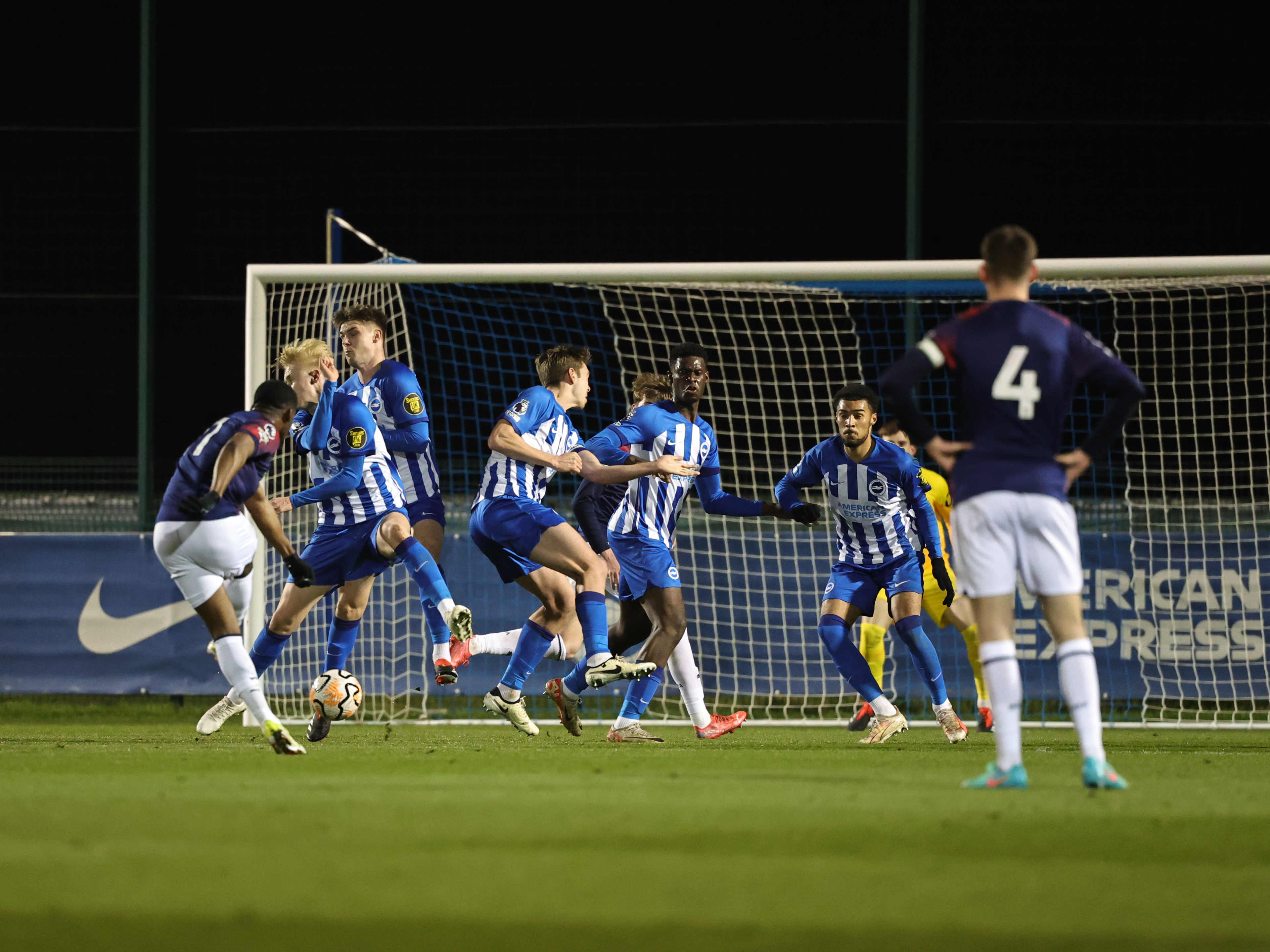 A photo of Jovan Malcolm, in the 23/24 red and blue away colours, shooting a free-kick into a Brighton wall in an U21s match at Brighton's training ground