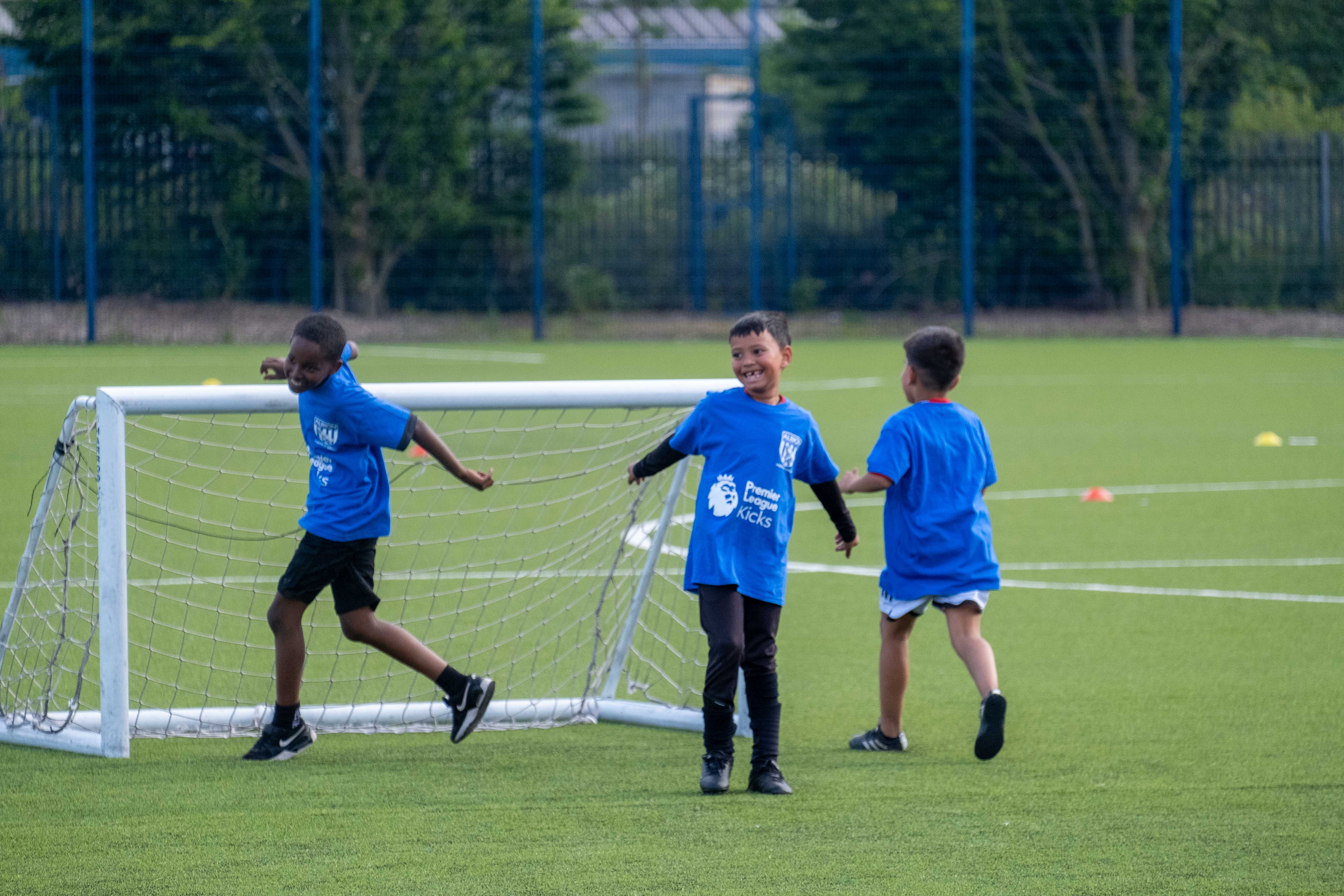 Three players wearing blue bibs smile and laugh during the South Asian Emerging Talent Festival on the Academy pitch.