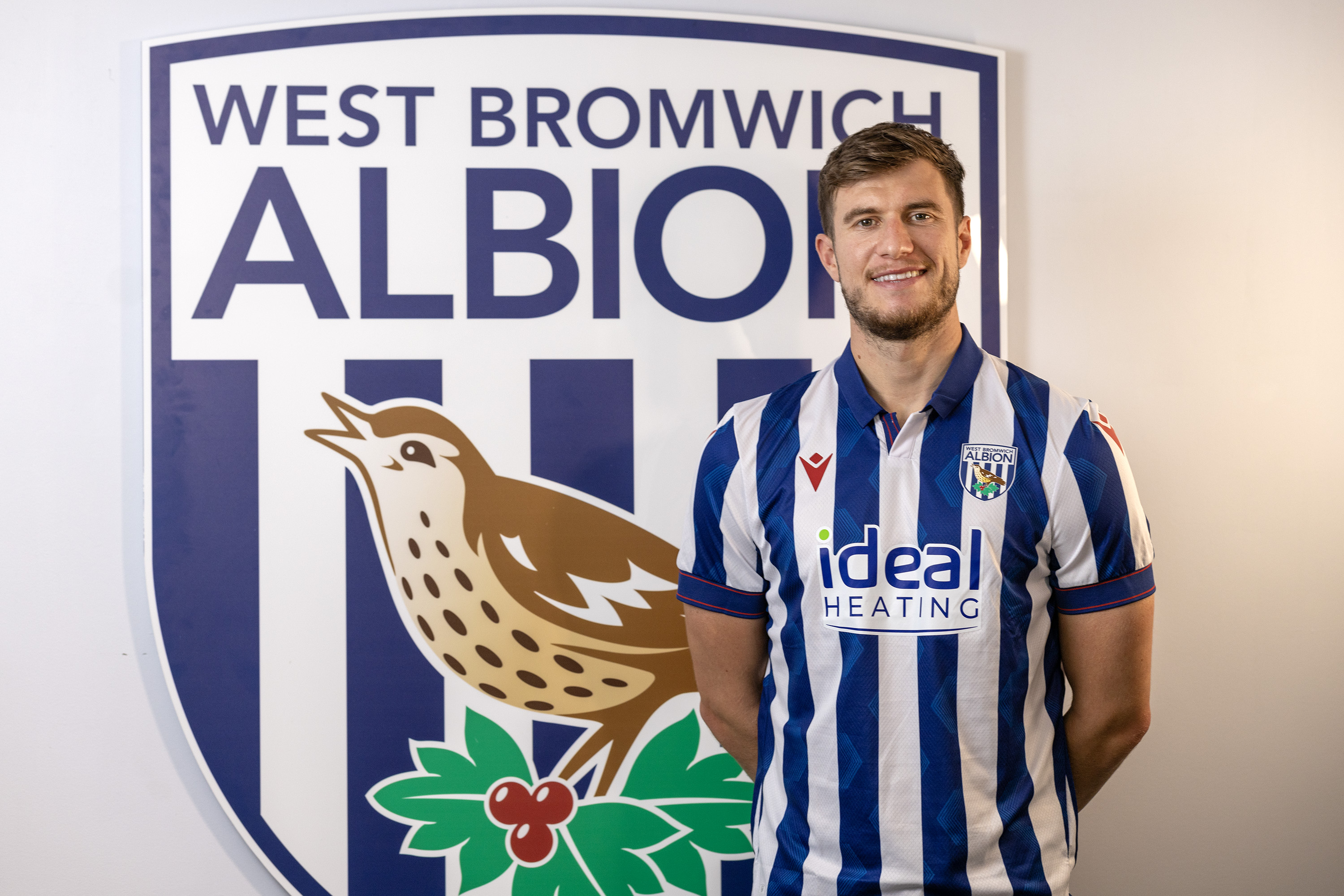 Paddy McNair smiling at the camera while stood in a home shirt in front of an Albion badge 