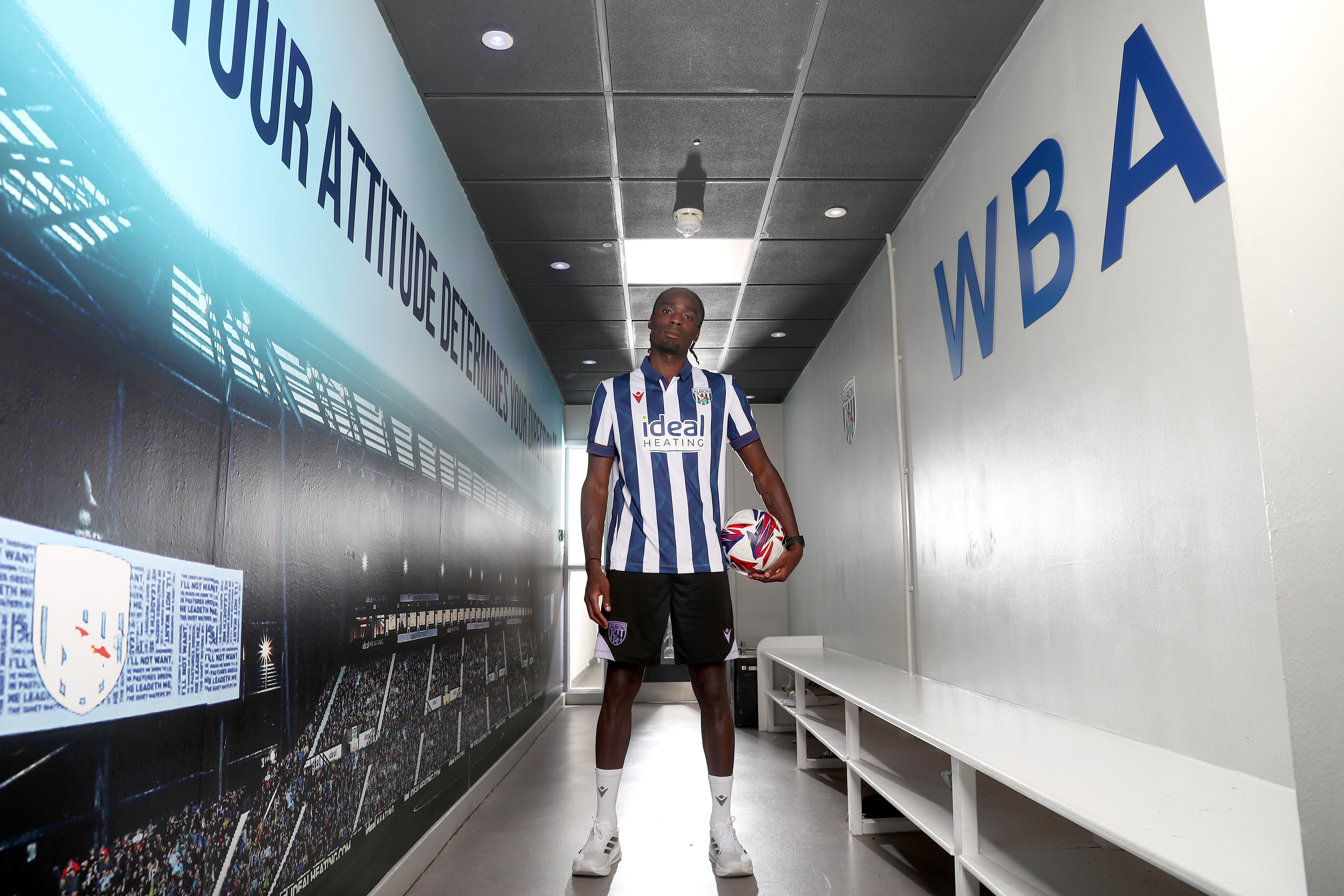 Devante Cole staring at the camera in a corridor holding a ball and wearing a home shirt 