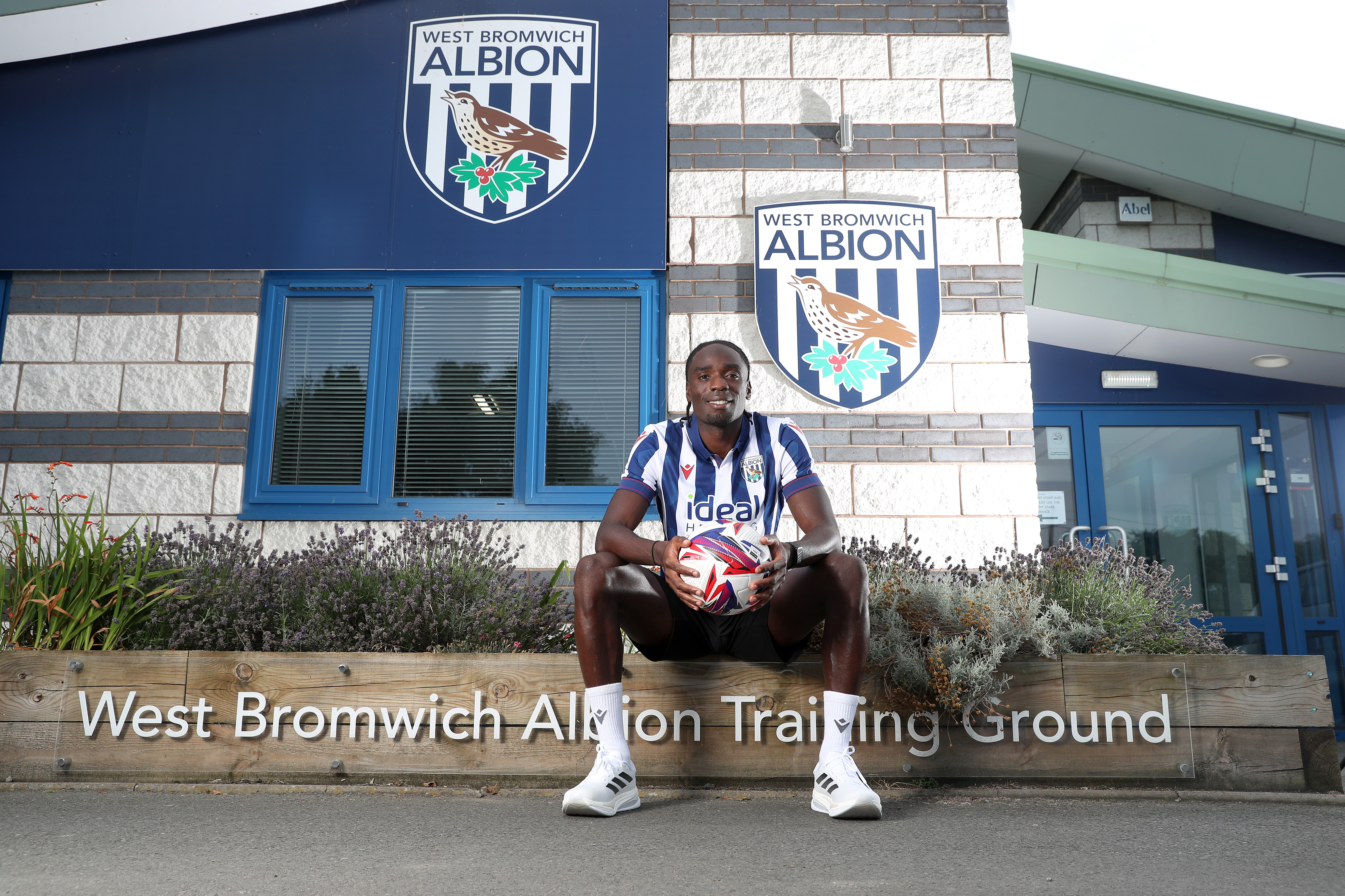 Devante Cole sat outside the training ground entrance holding a ball and wearing a home shirt 