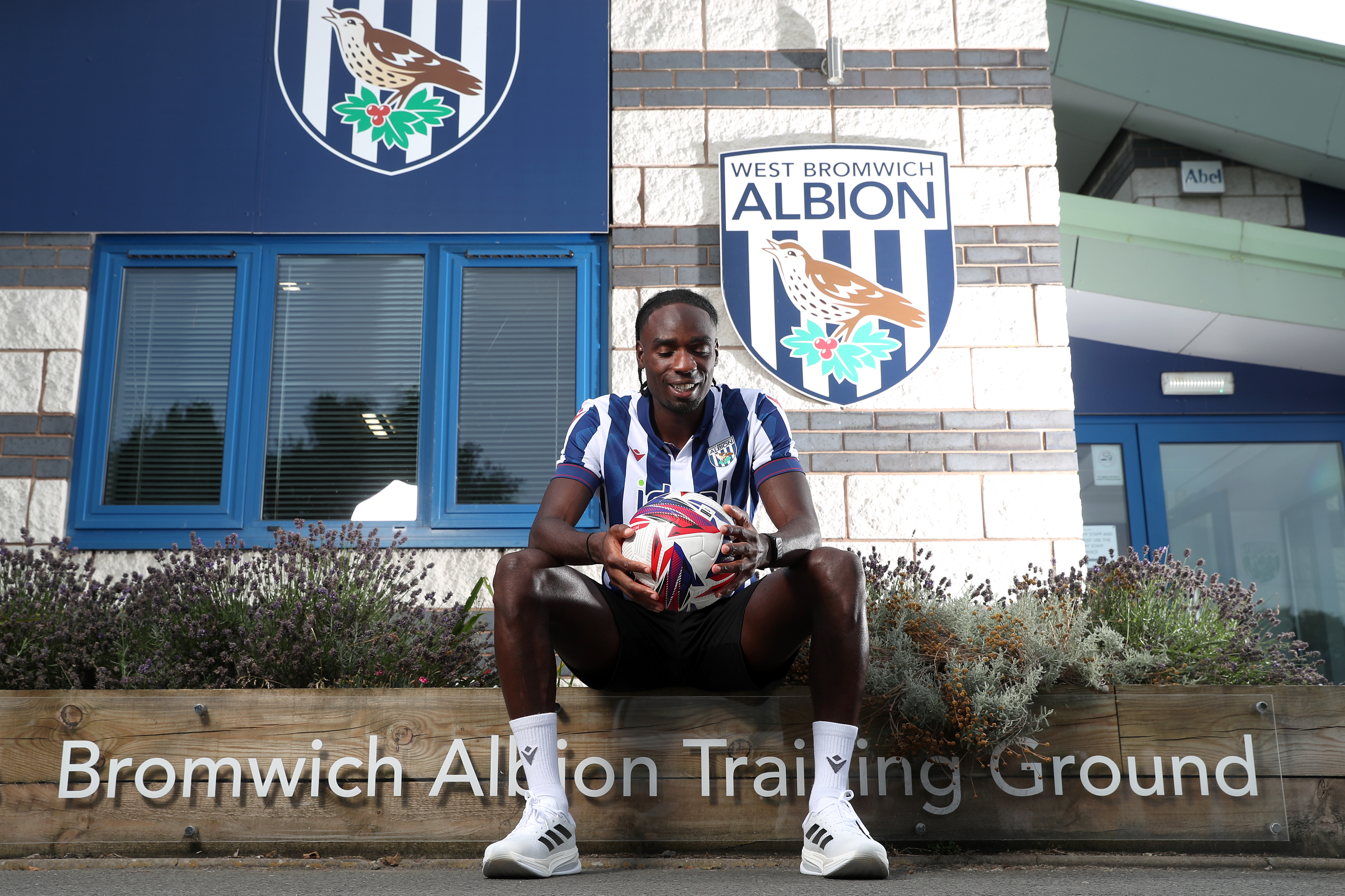 Devante Cole smiling while looking down at a ball sat outside the training ground entrance in a home shirt 