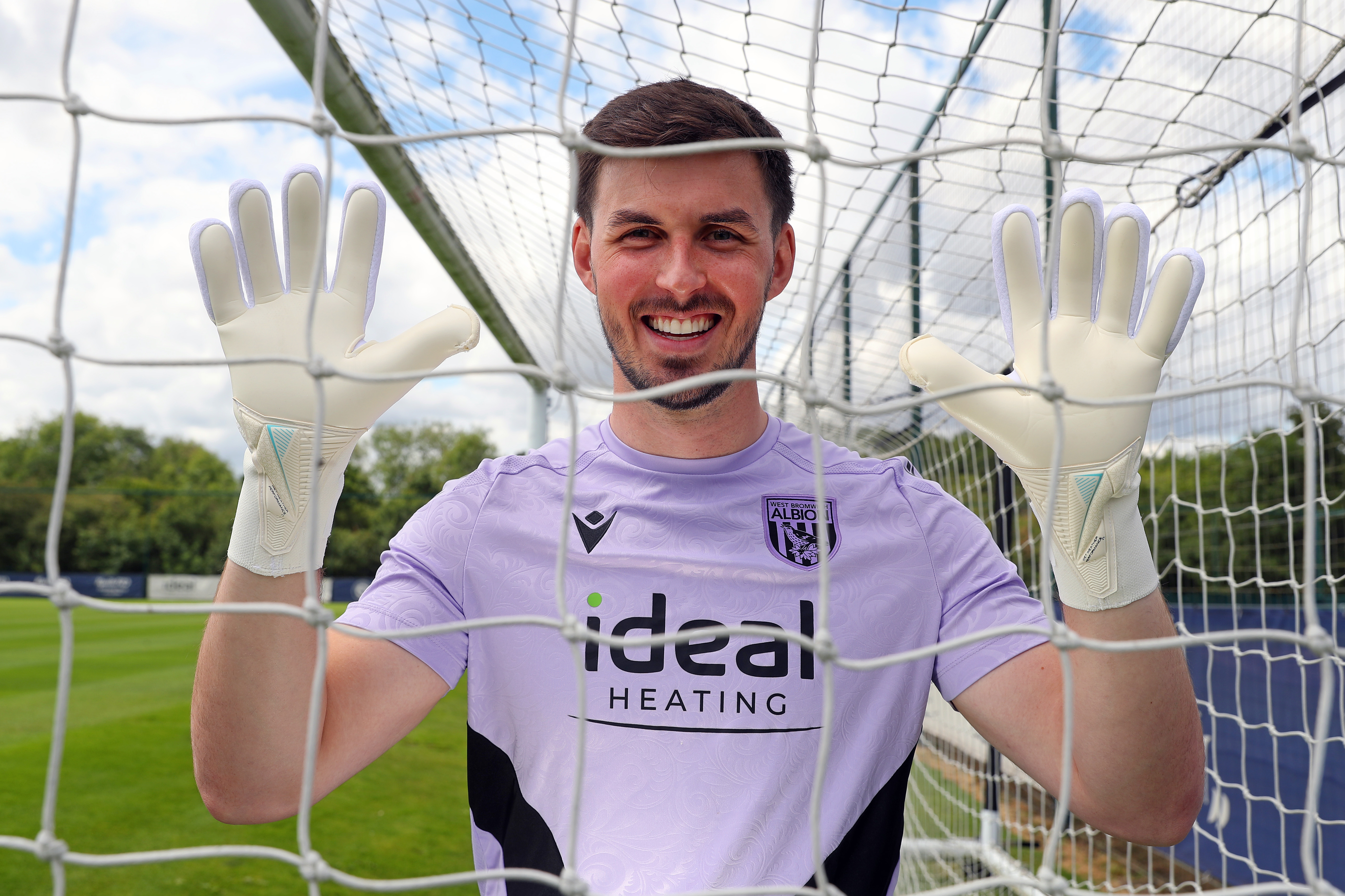 Joe Wildsmith smiling at the camera while holding his gloves up and looking through a goal net