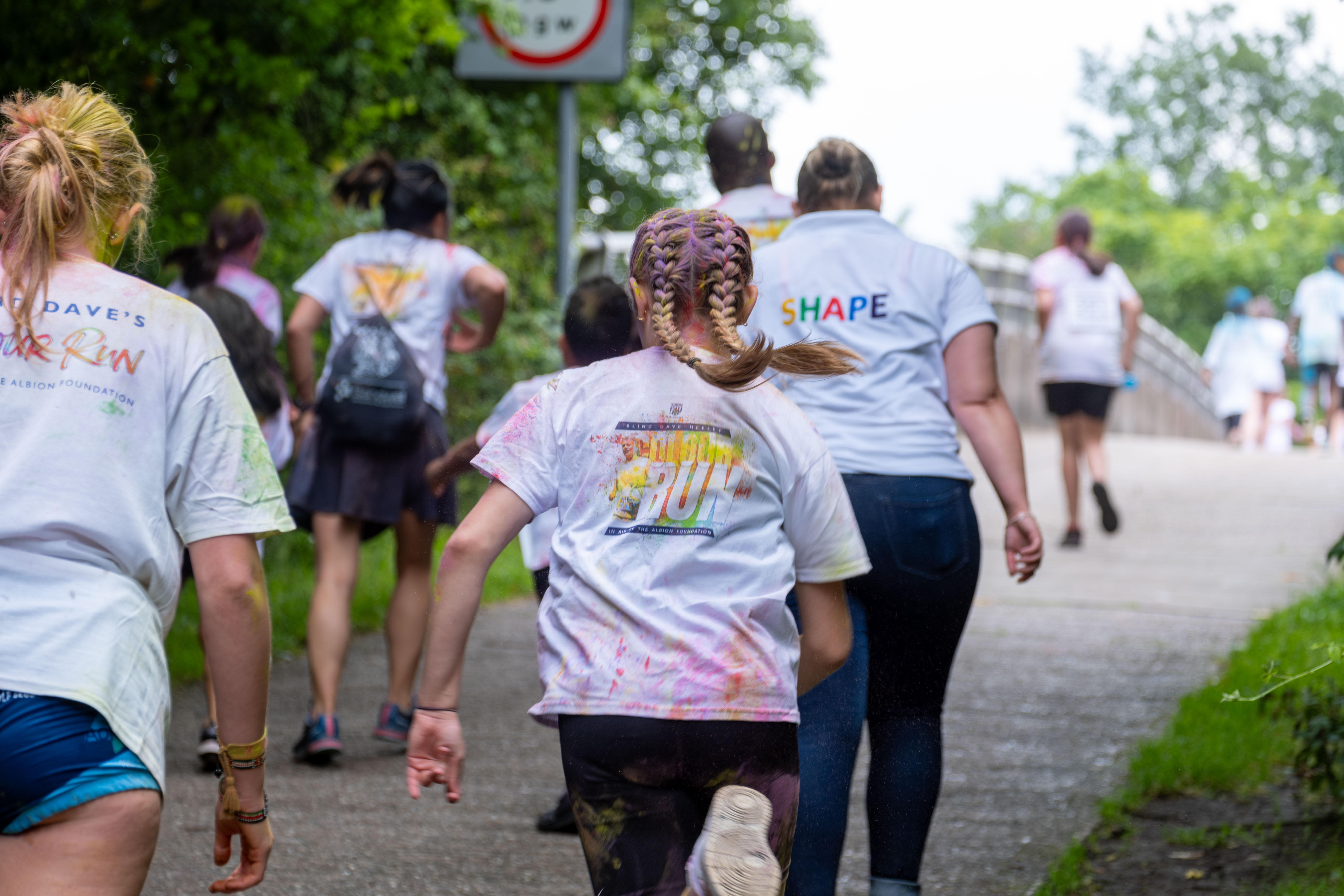 Back of Runner heading town motorway bridge, with Colour Run branding on display & SHAPE Festival displayed on one person back.