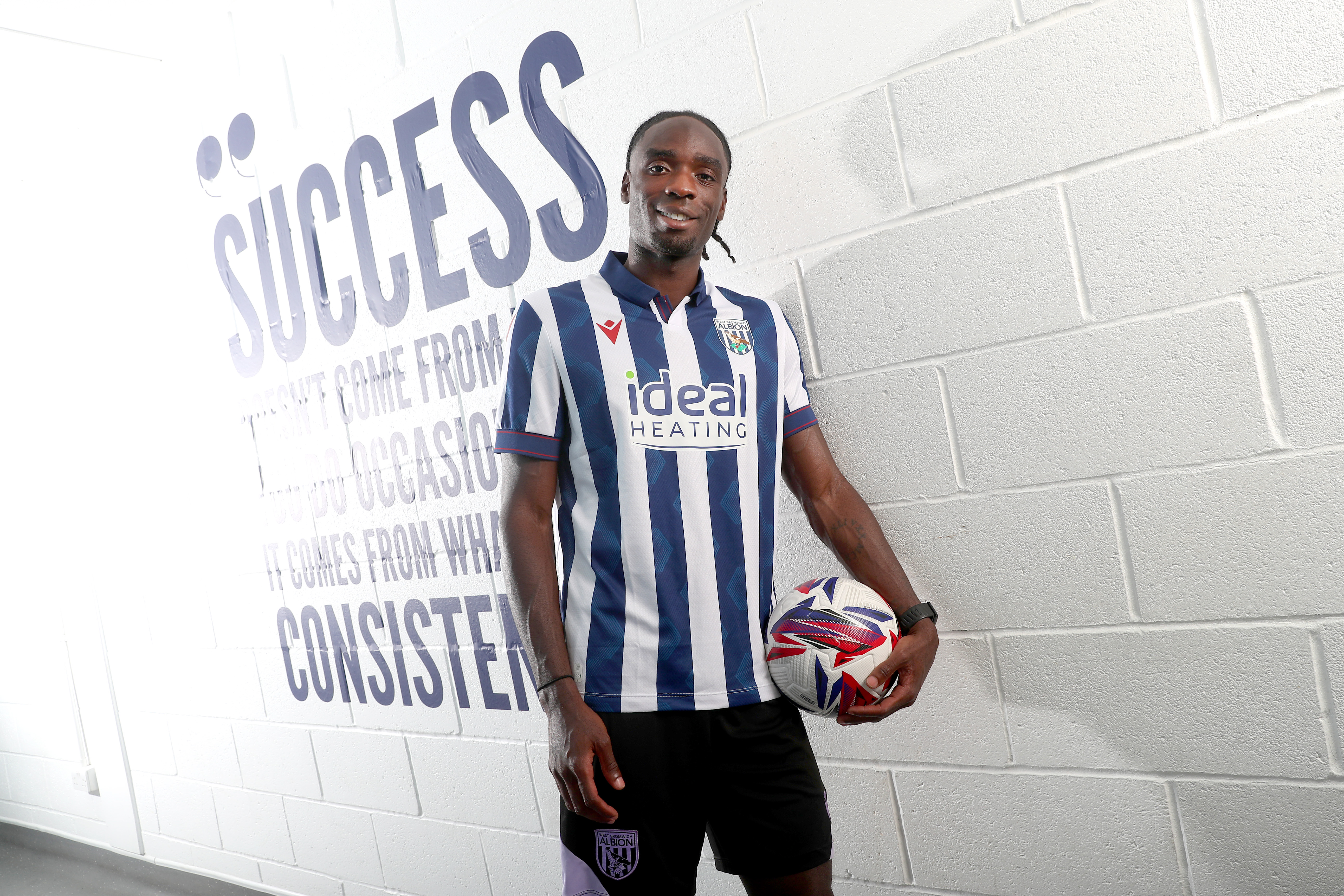 Devante Cole leaning against a wall with the word success written on it while smiling at the camera holding a football and wearing the home kit 