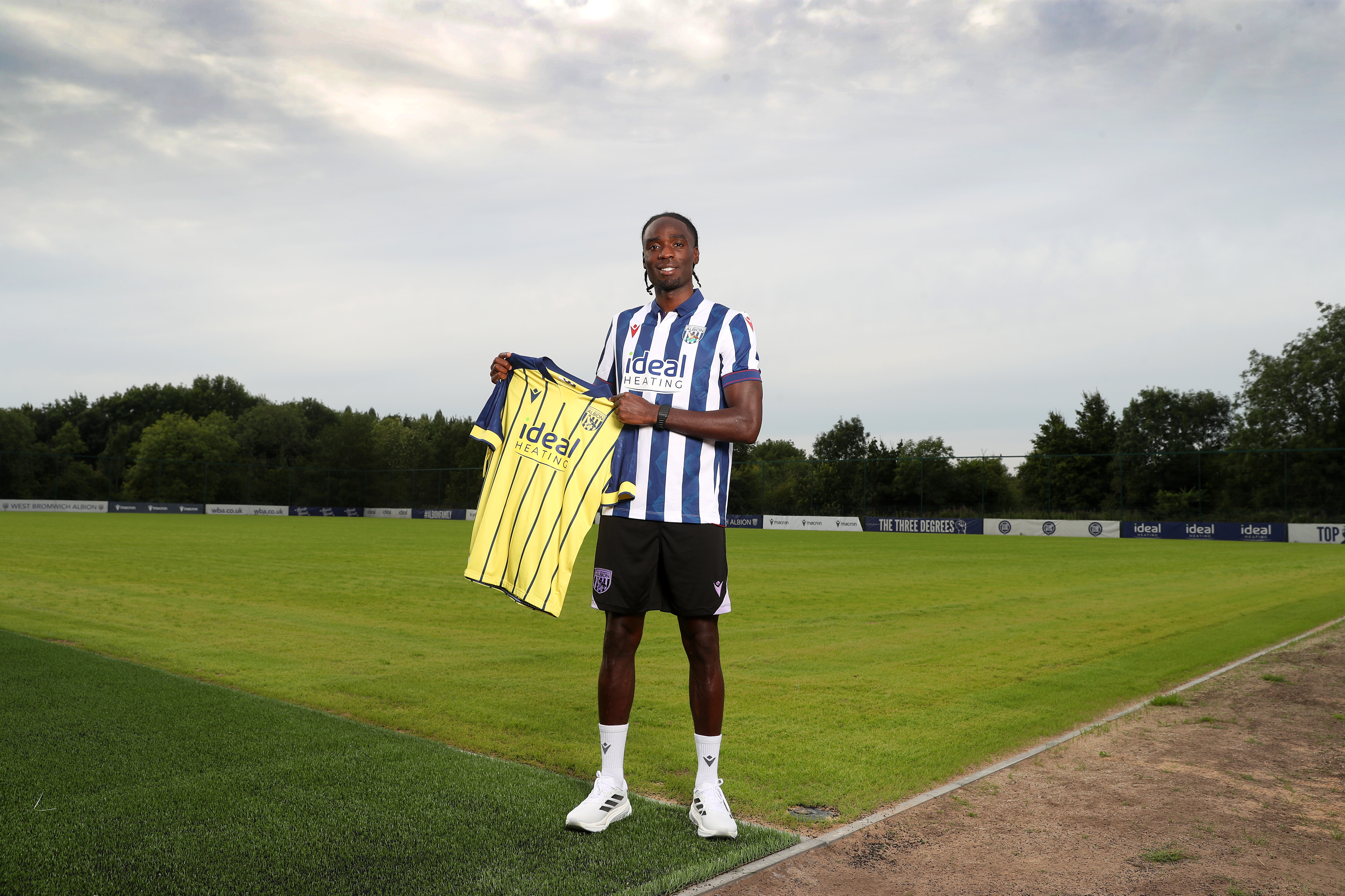 Devante Cole smiling at the camera while wearing a home shirt and holding up a yellow away shirt 