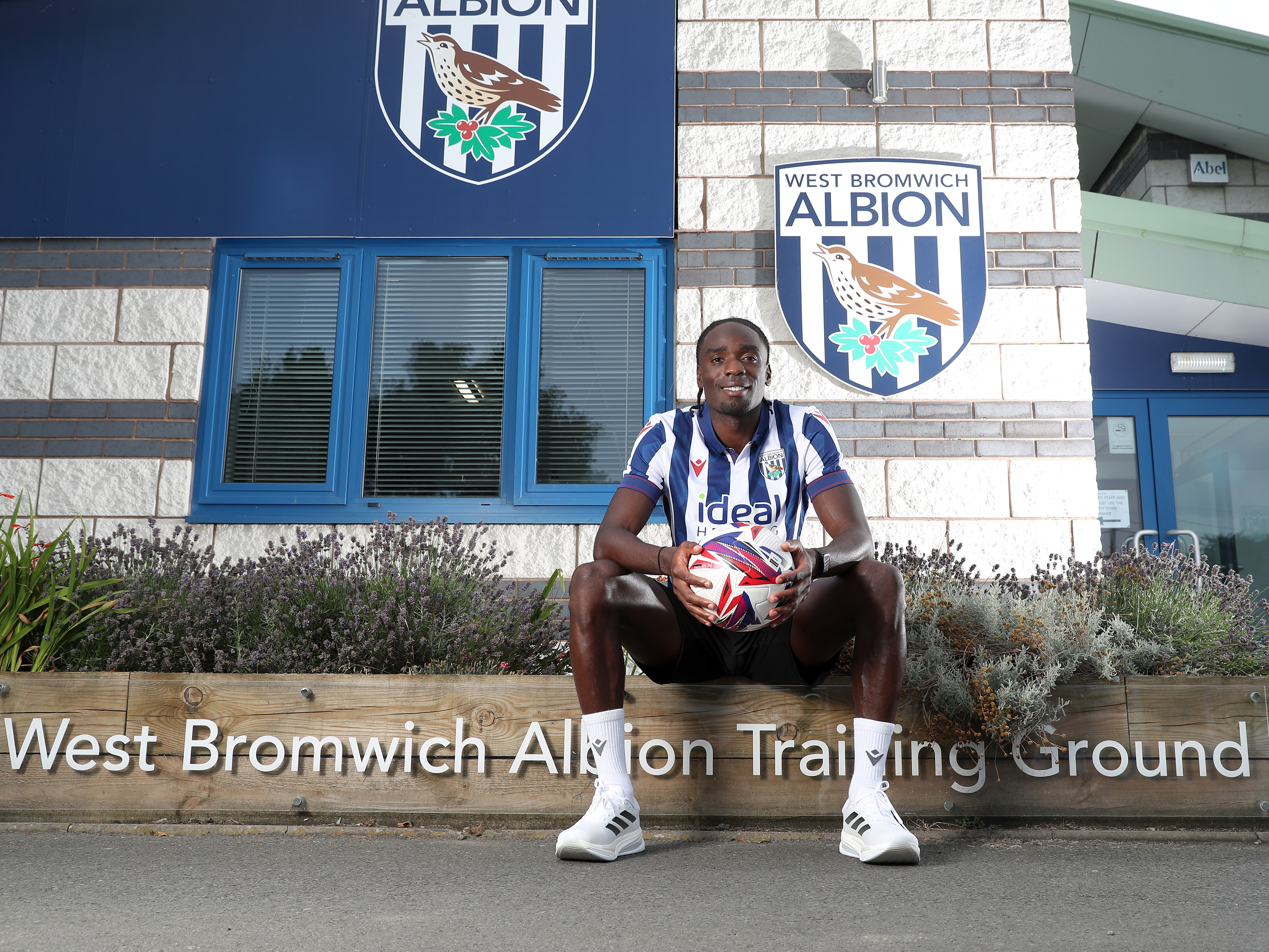 Devante Cole sat outside the entrance of the training ground holding a ball while wearing a home shirt and smiling at the camera 