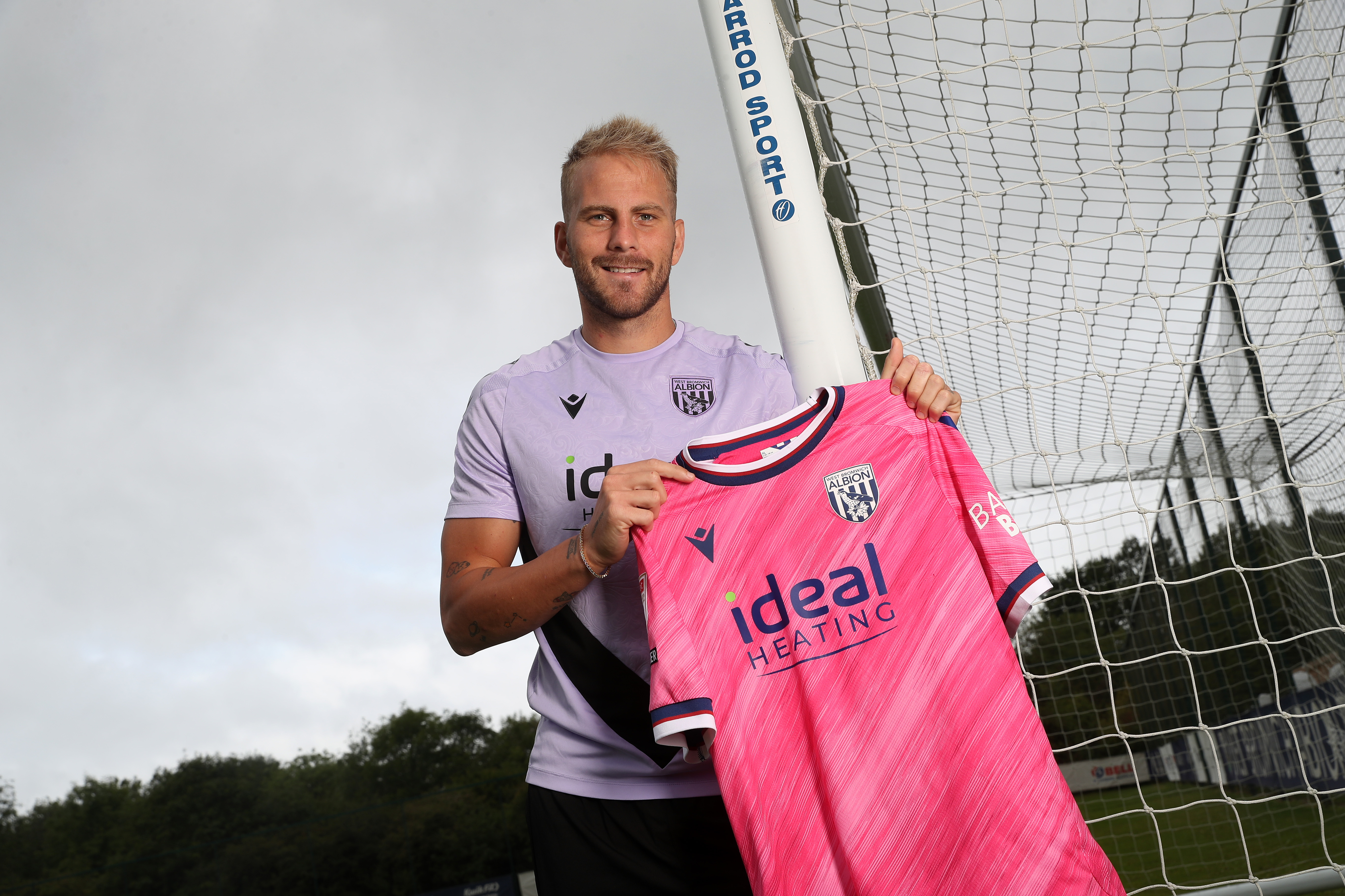 Uroš Račić smiling at the camera while stood holding up a pink away shirt 