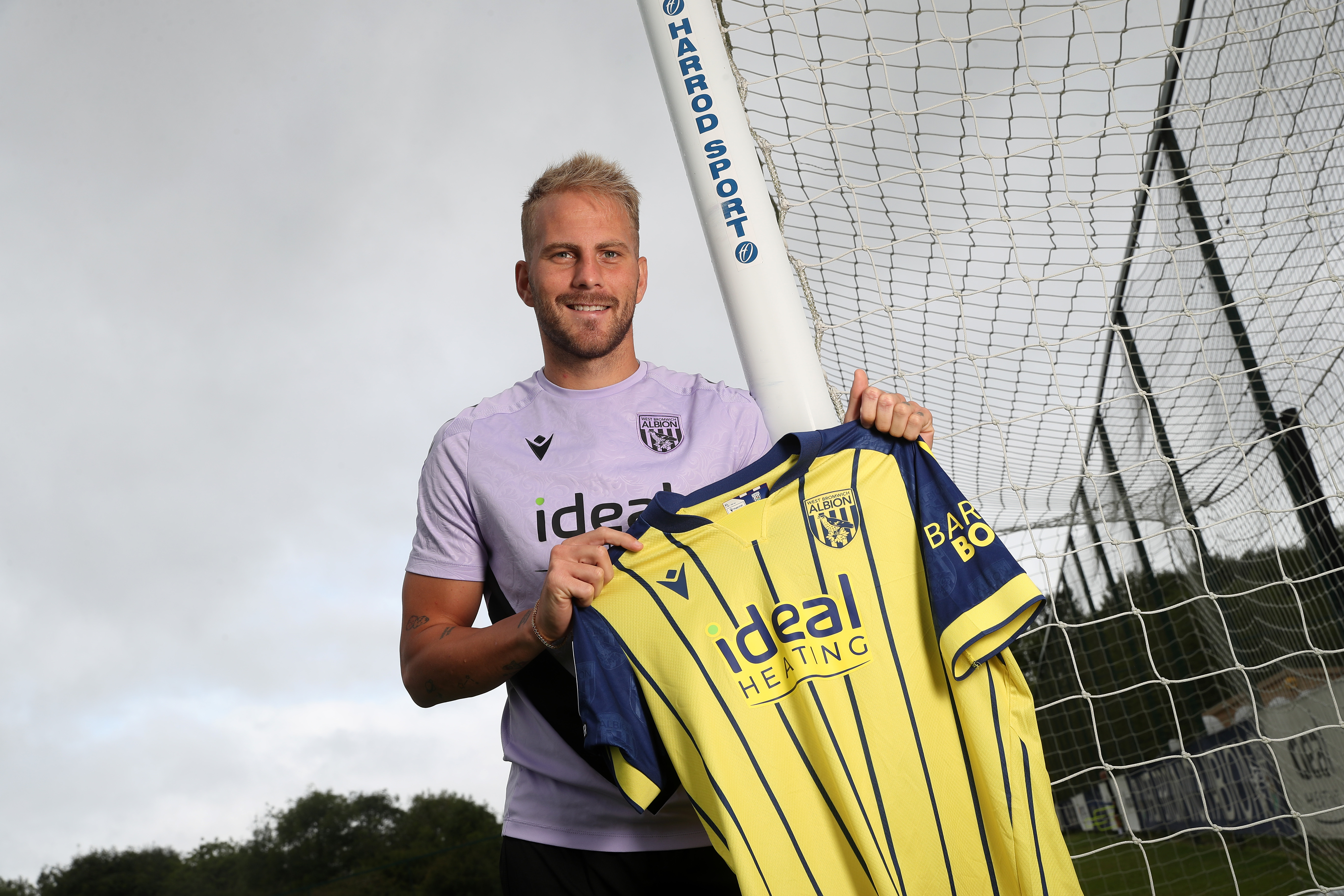 Uroš Račić smiling at the camera while stood holding up a yellow away shirt 