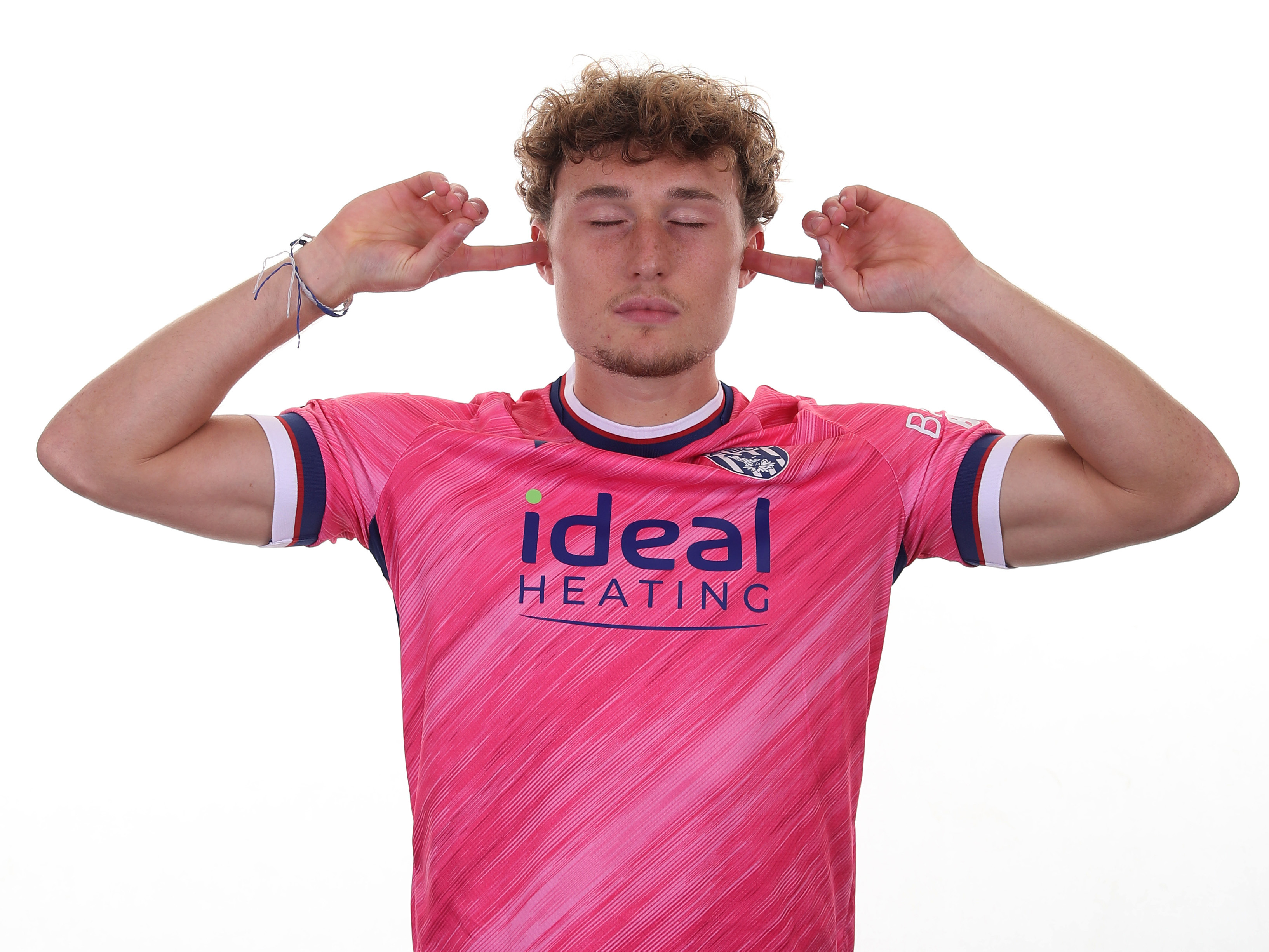 Callum Styles with his fingers in his ears and his eyes closed while stood in the pink away shirt 