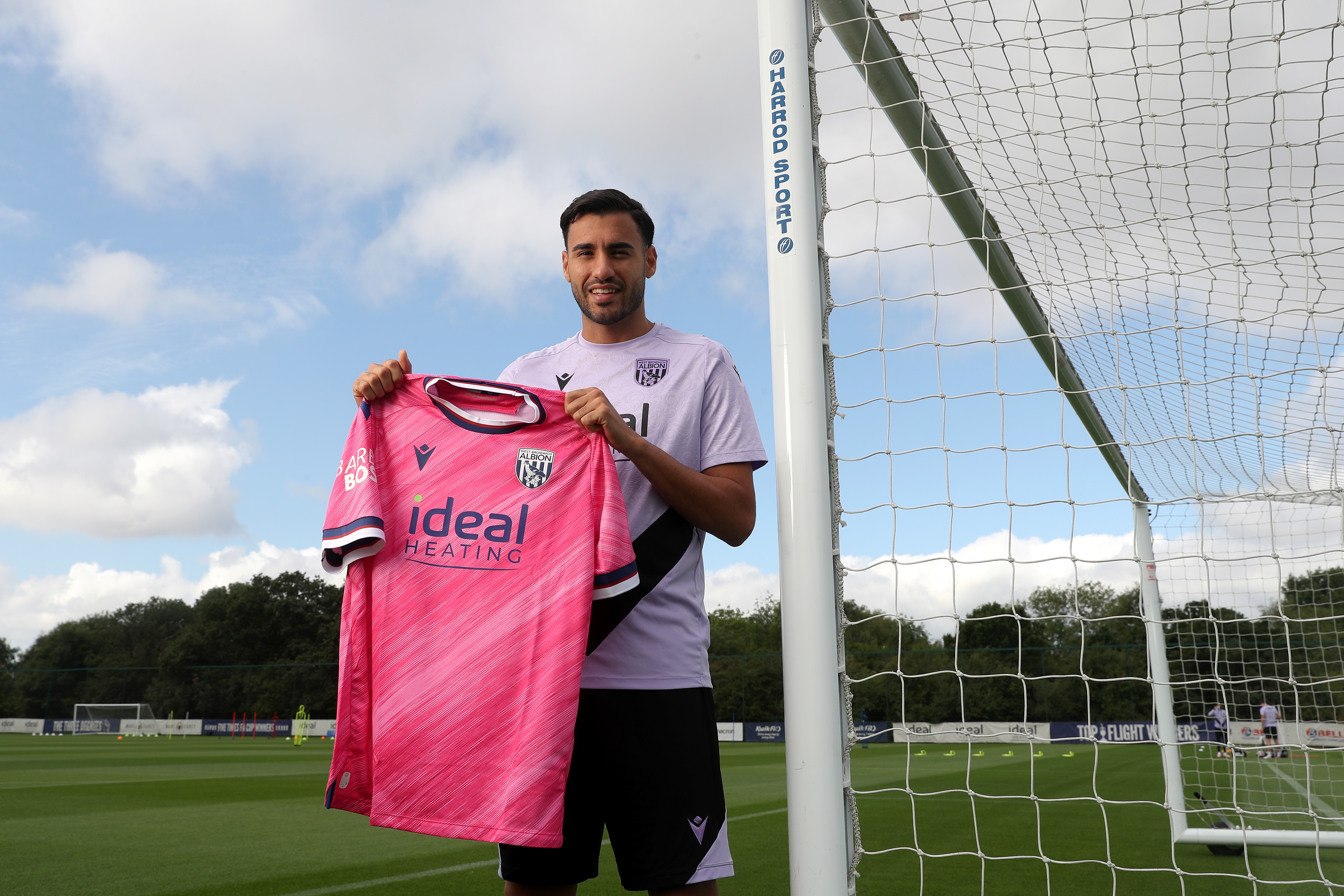 Gianluca Frabotta smiling at the camera while stood against a goal post holding up a pink away shirt