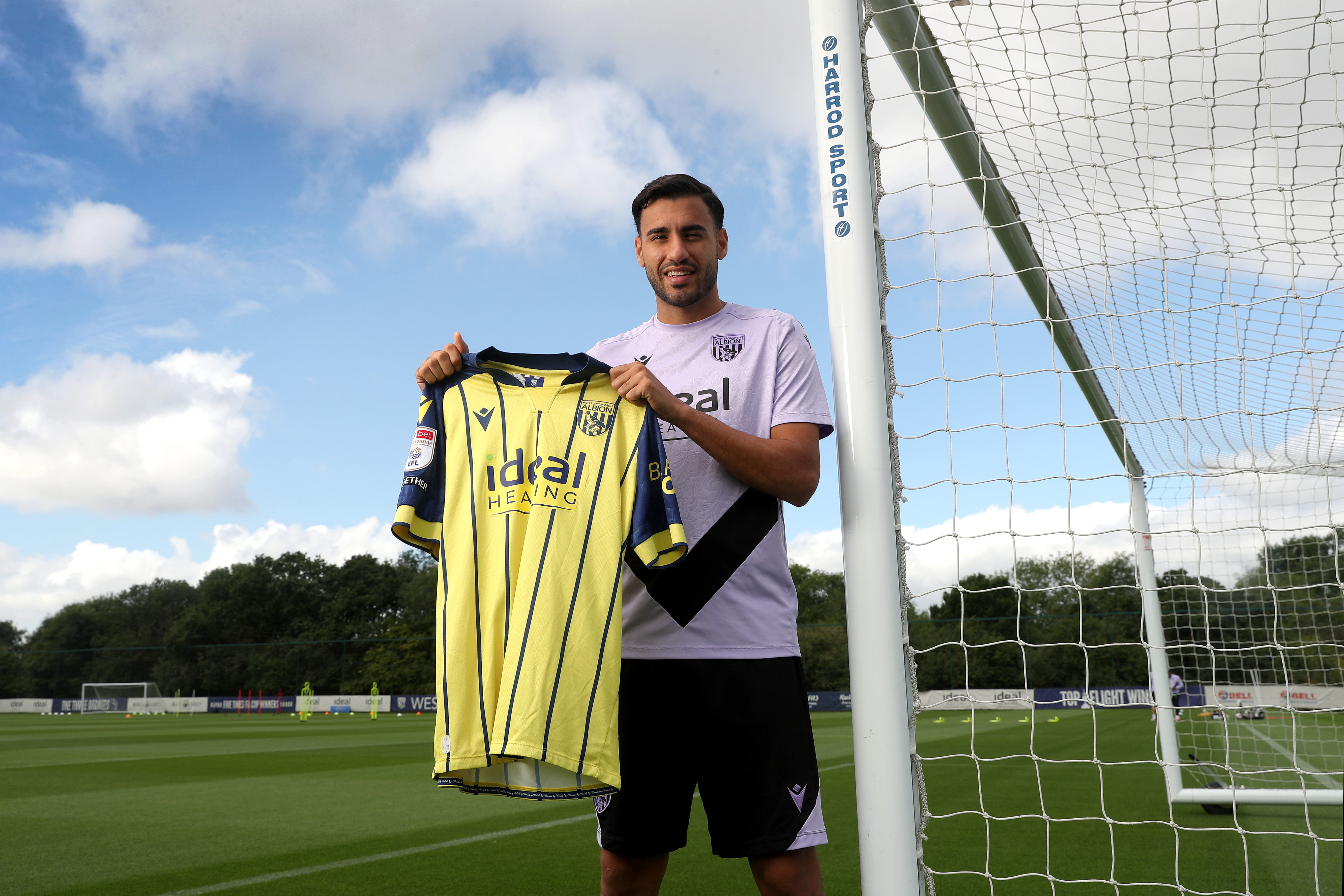 Gianluca Frabotta smiling at the camera while stood against a goal post holding up a yellow away shirt