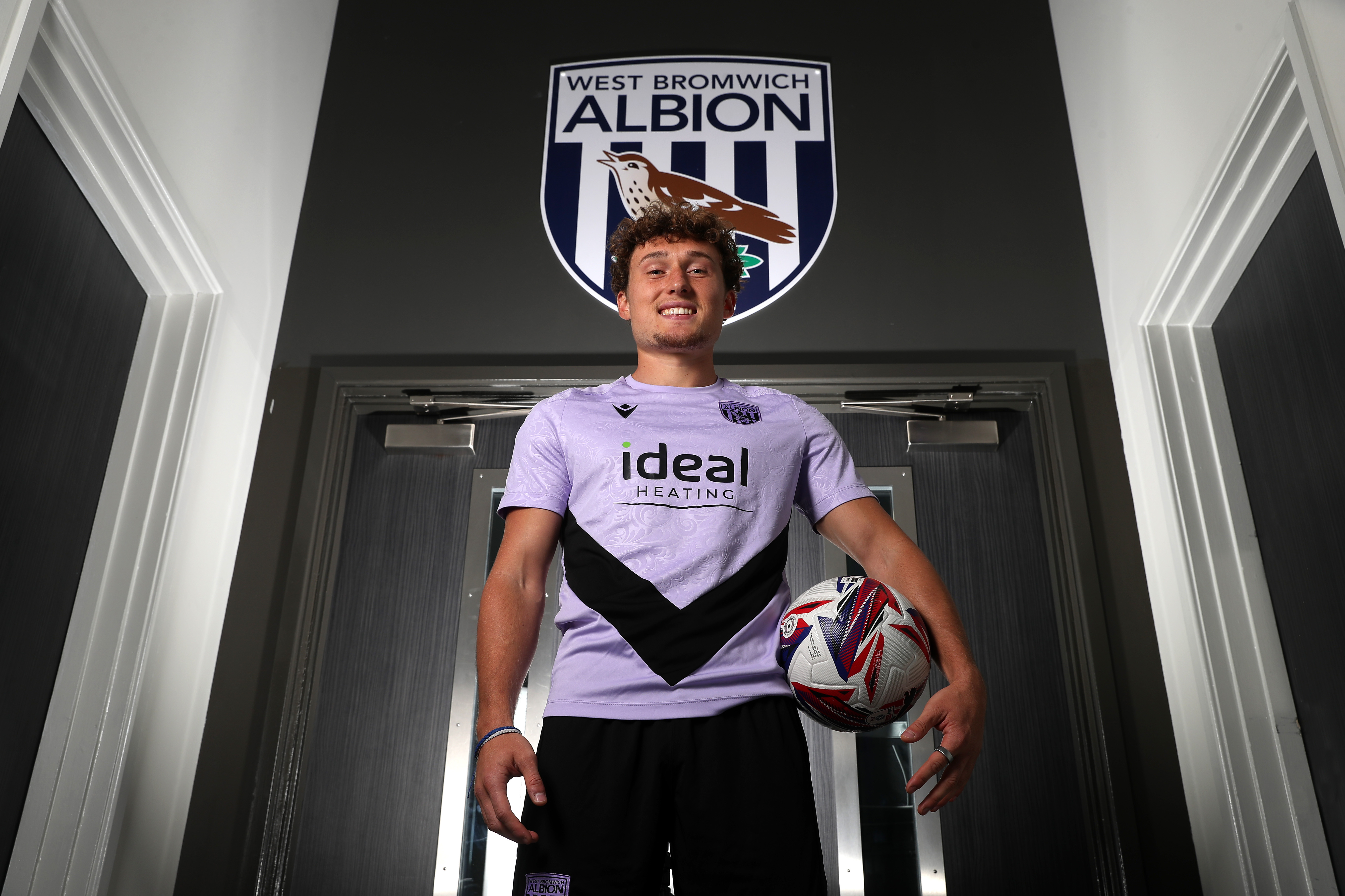 Callum Styles smiling at the camera while holding a ball while stood in a corridor 