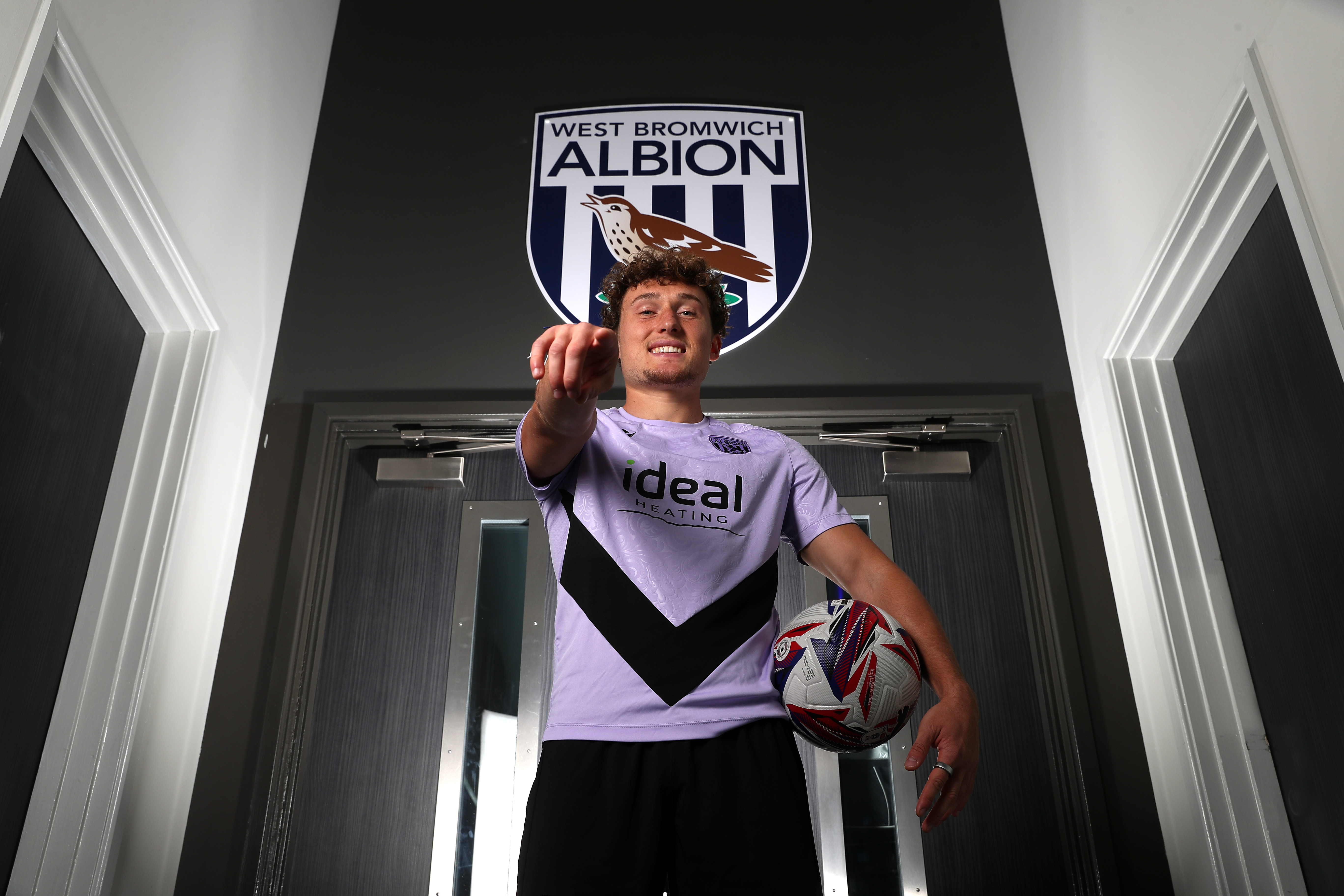 Callum Styles smiling at the camera while holding a ball and pointing at the camera in a corridor 