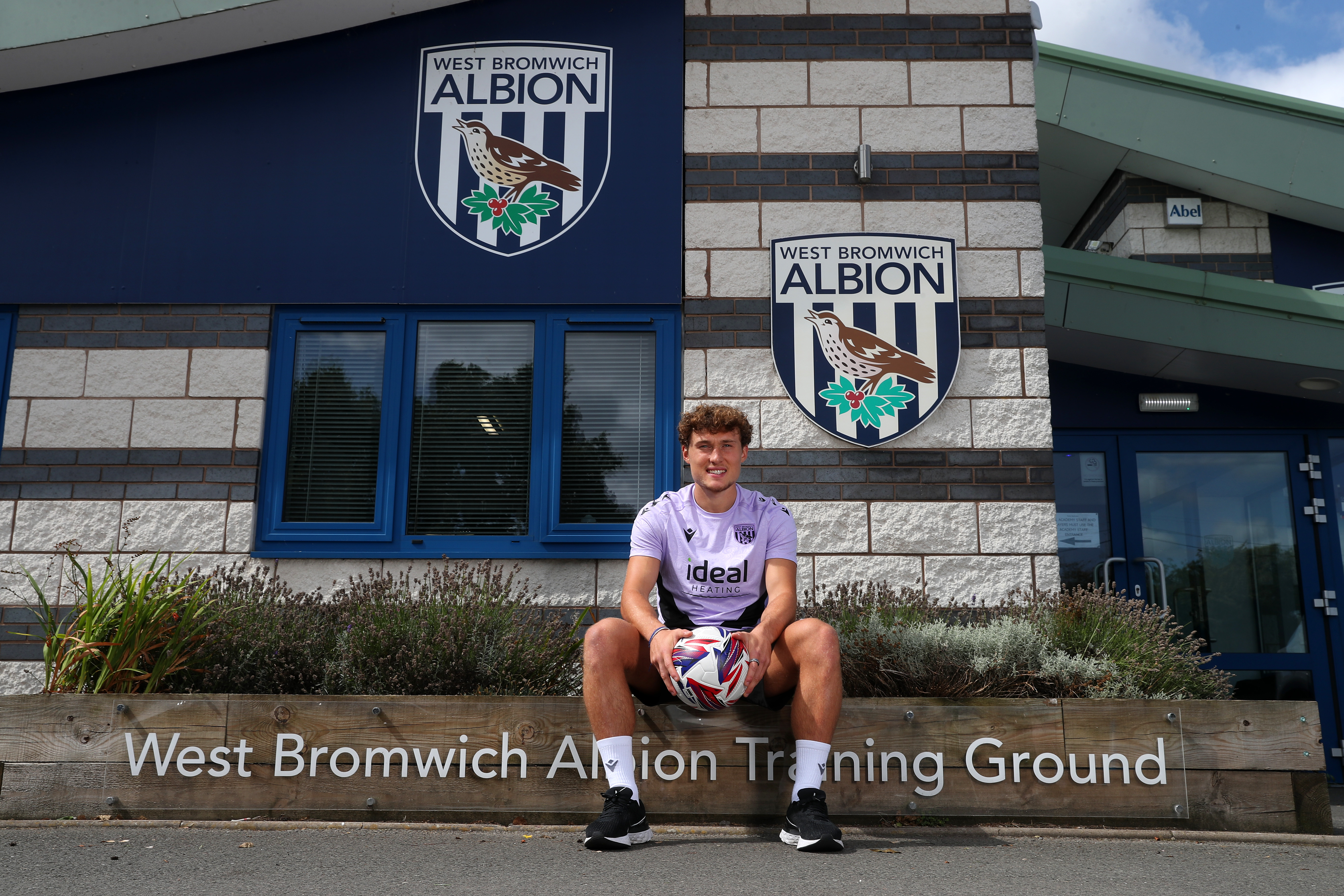 Callum Styles smiling at the camera while holding a ball sat outside the front of the training ground 