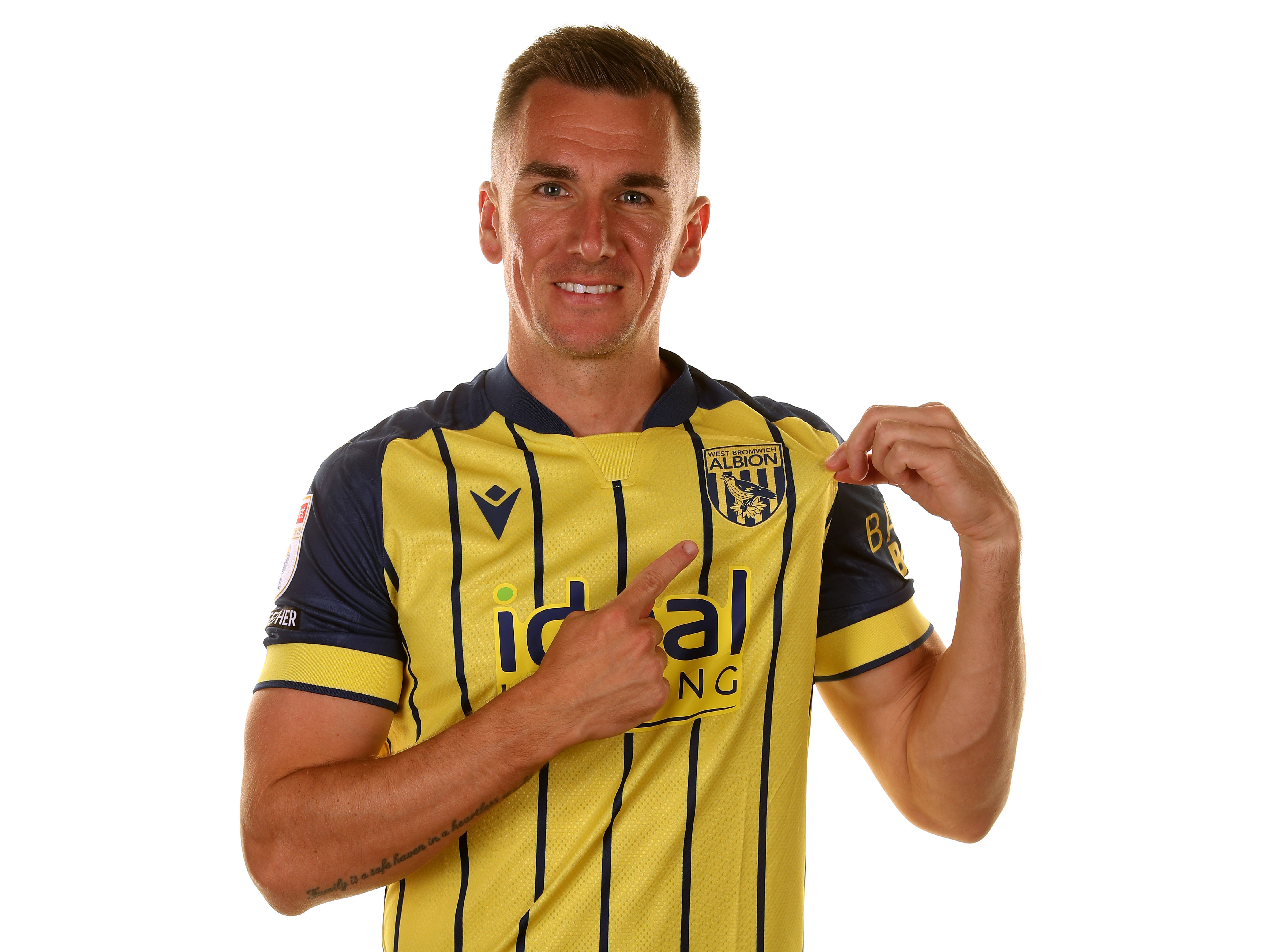 Jed Wallace pointing to the badge smiling at the camera wearing the yellow away shirt 