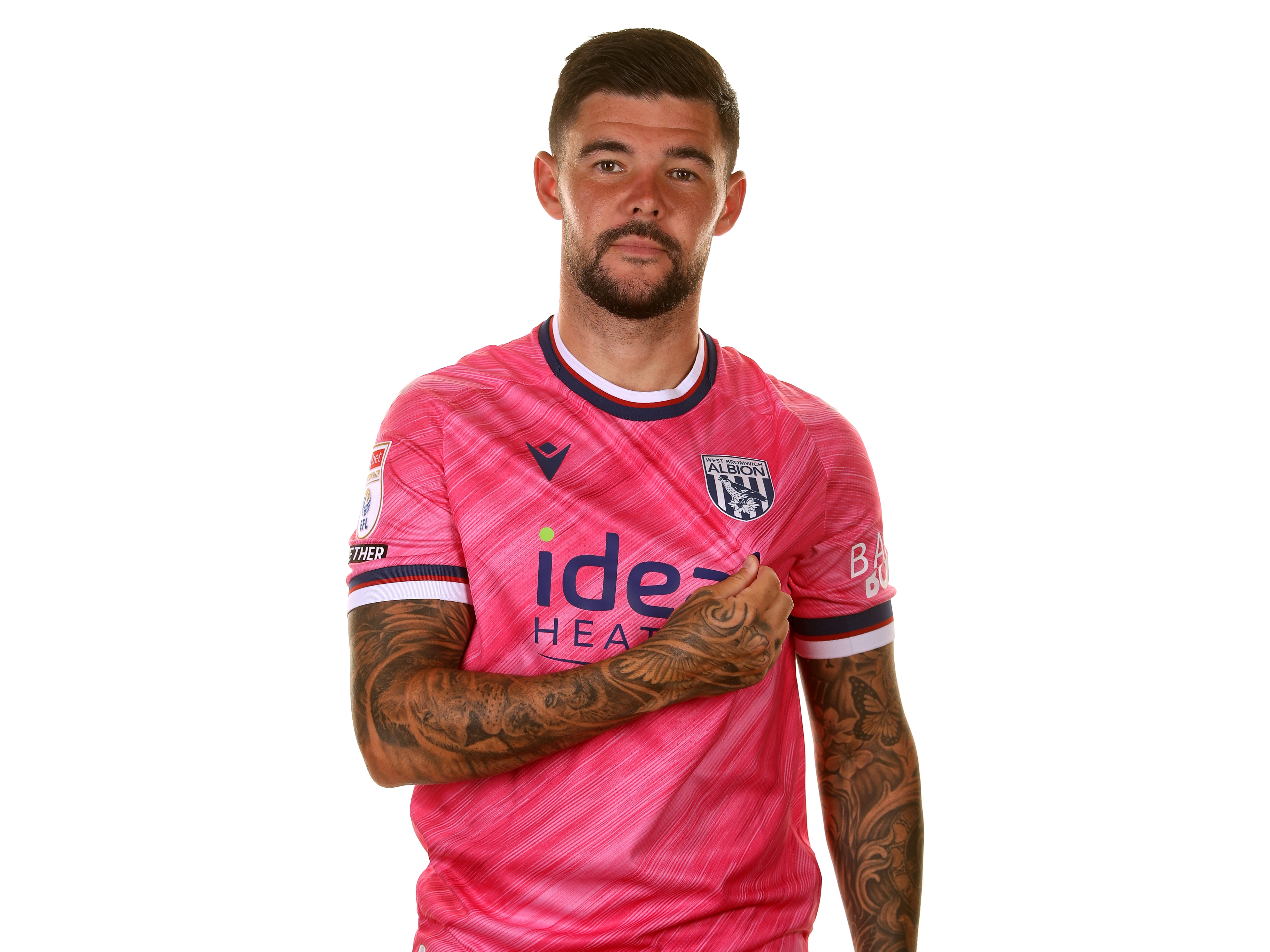 Alex Mowatt looking at the camera pulling on the badge while wearing the pink away shirt 