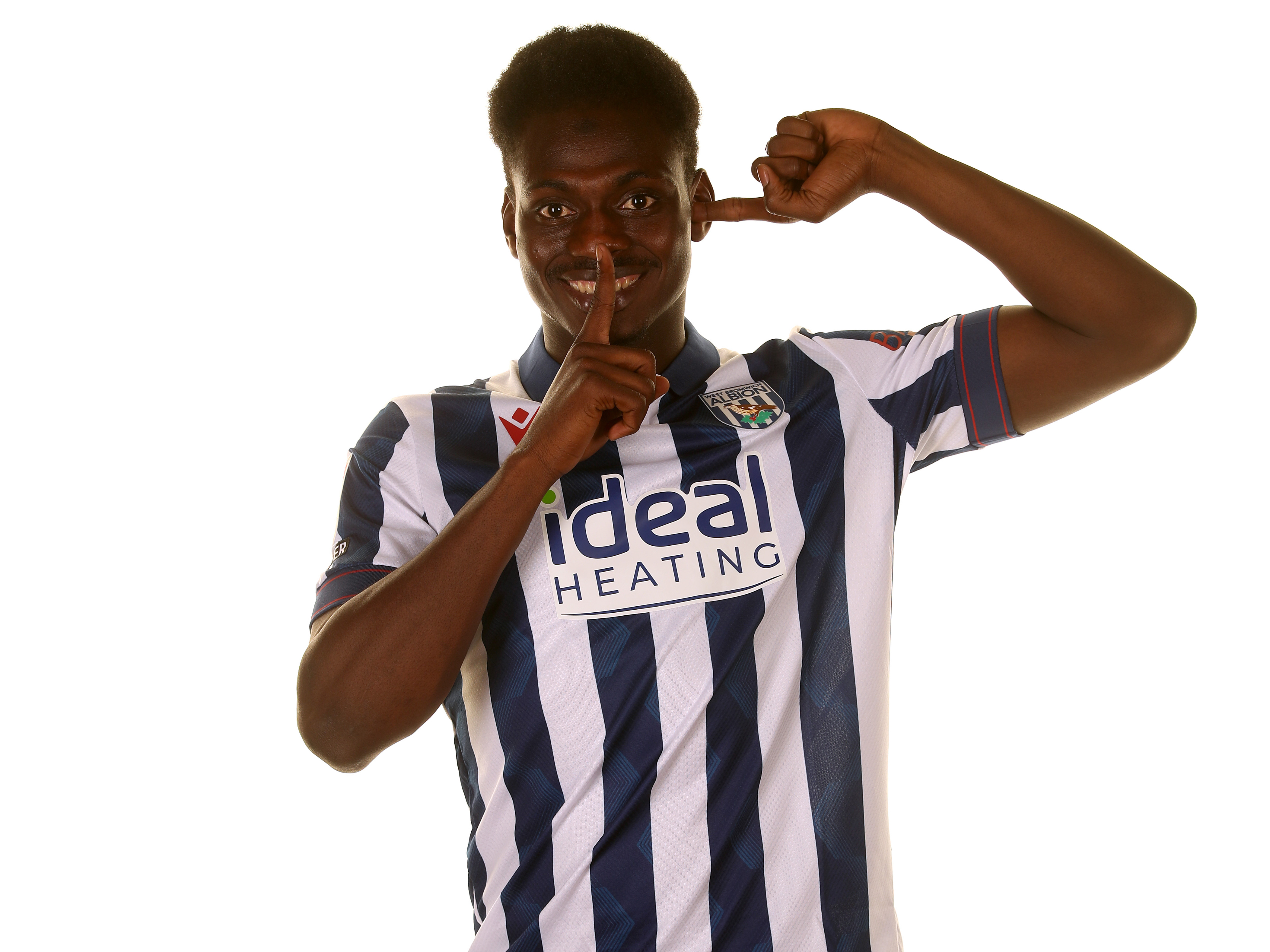 Ousmane Diakité holding one finger to his ear and another to his mouth smiling at the camera in a home shirt 