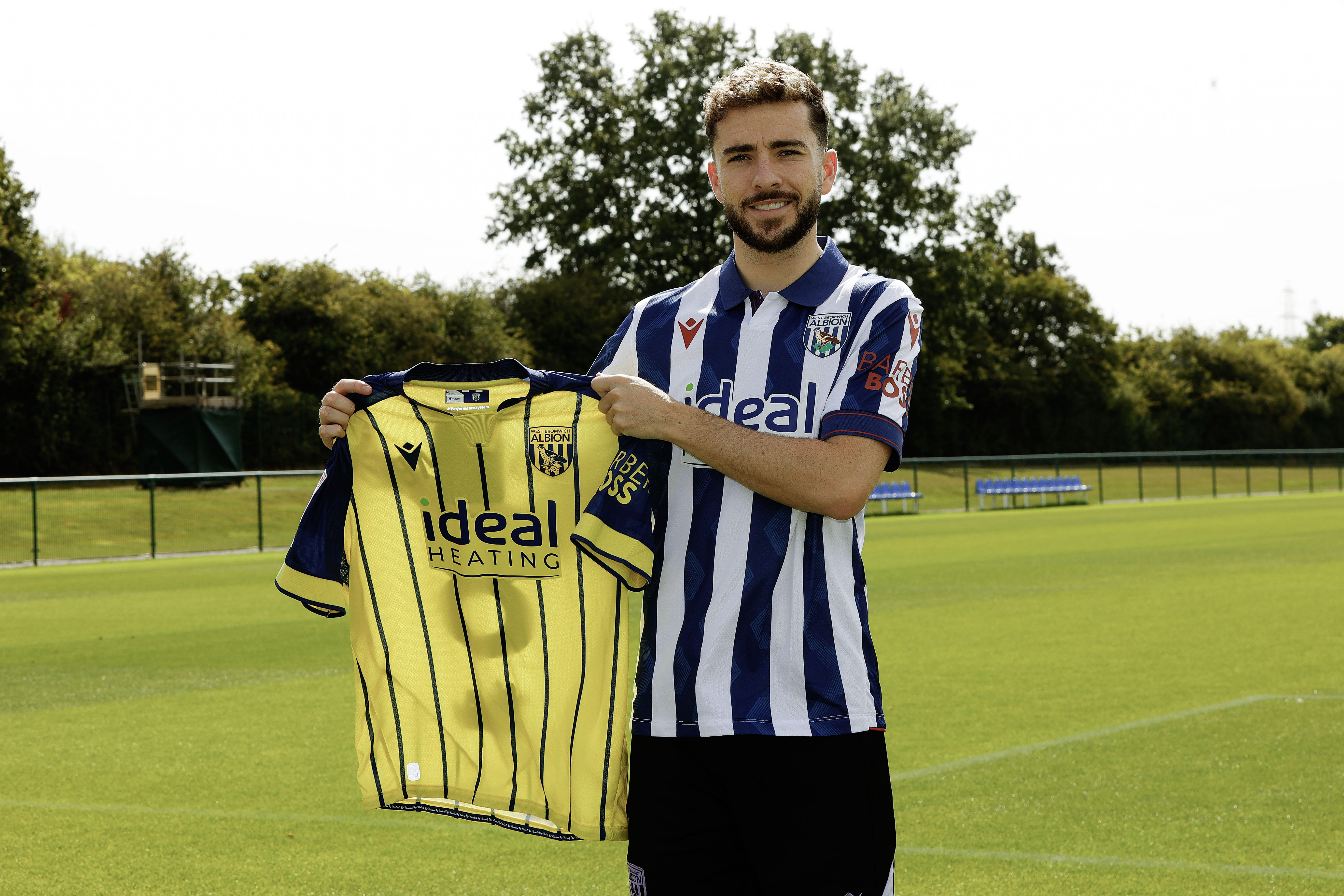 Mikey Johnston smiling at the camera holding up a yellow away shirt while stood in a home shirt 