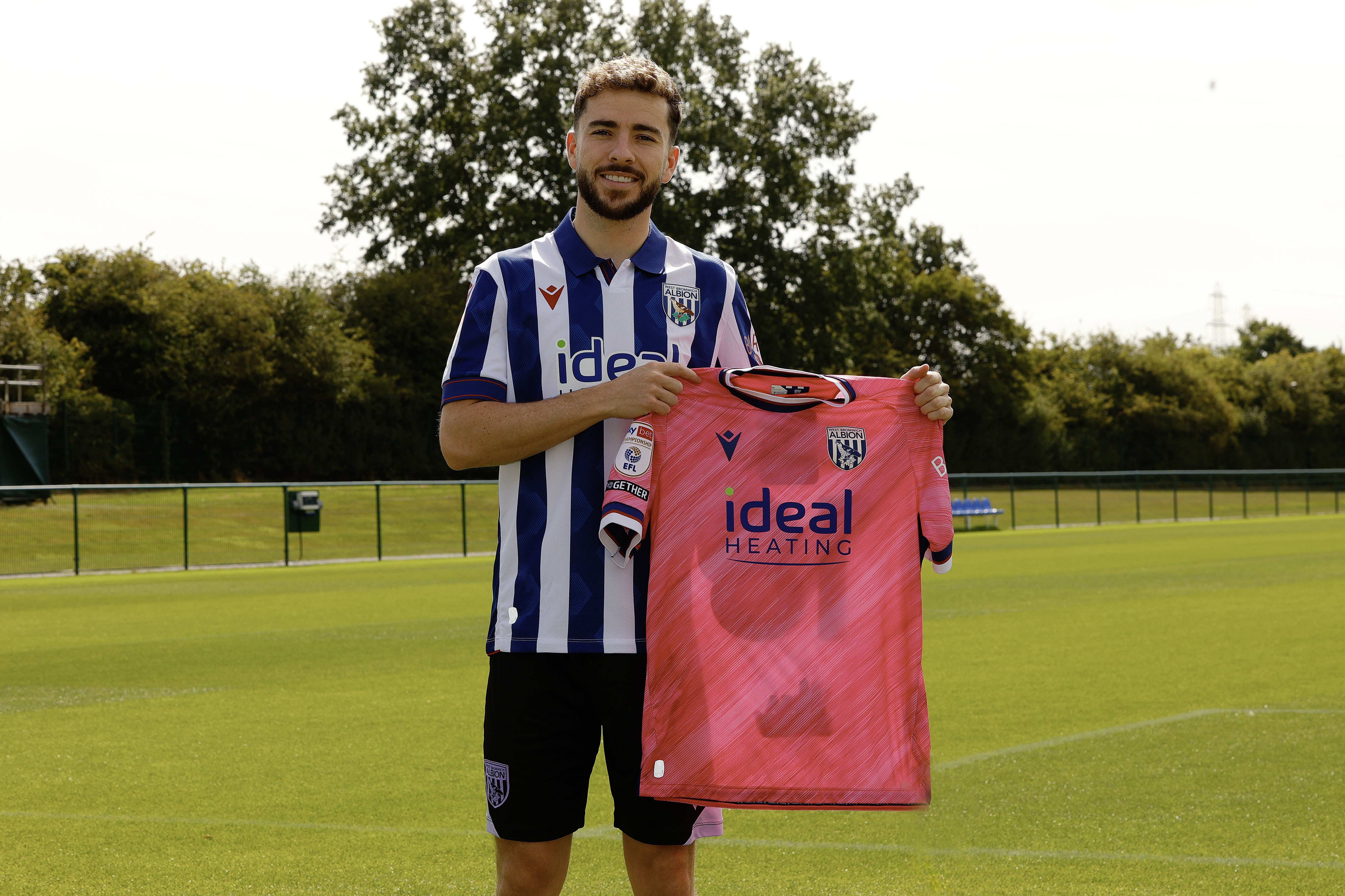 Mikey Johnston smiling at the camera holding up a pink away shirt while stood in a home shirt 