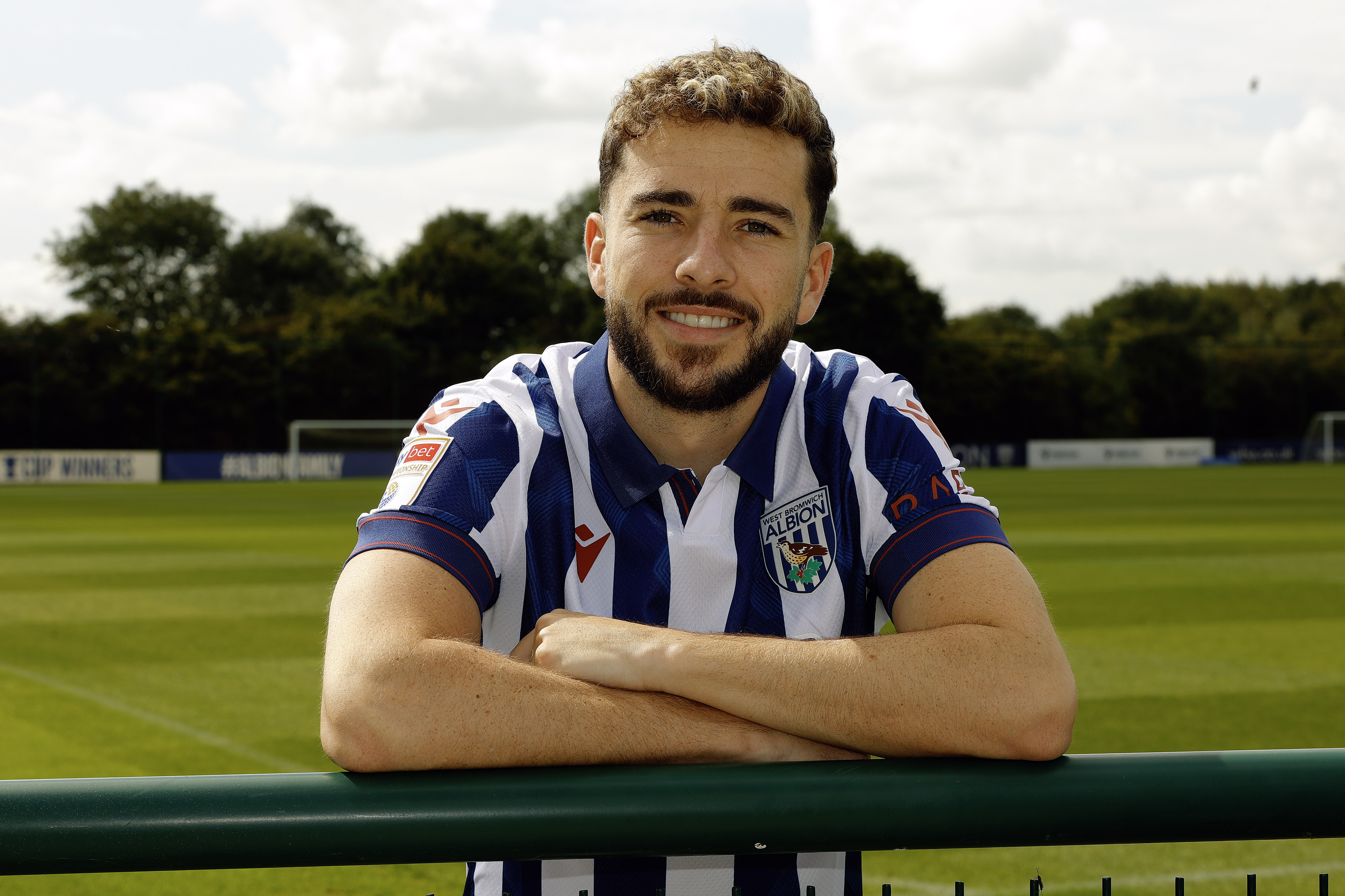 Mikey Johnston leaning over a barrier with his arms folded smiling at the camera while wearing a home shirt 