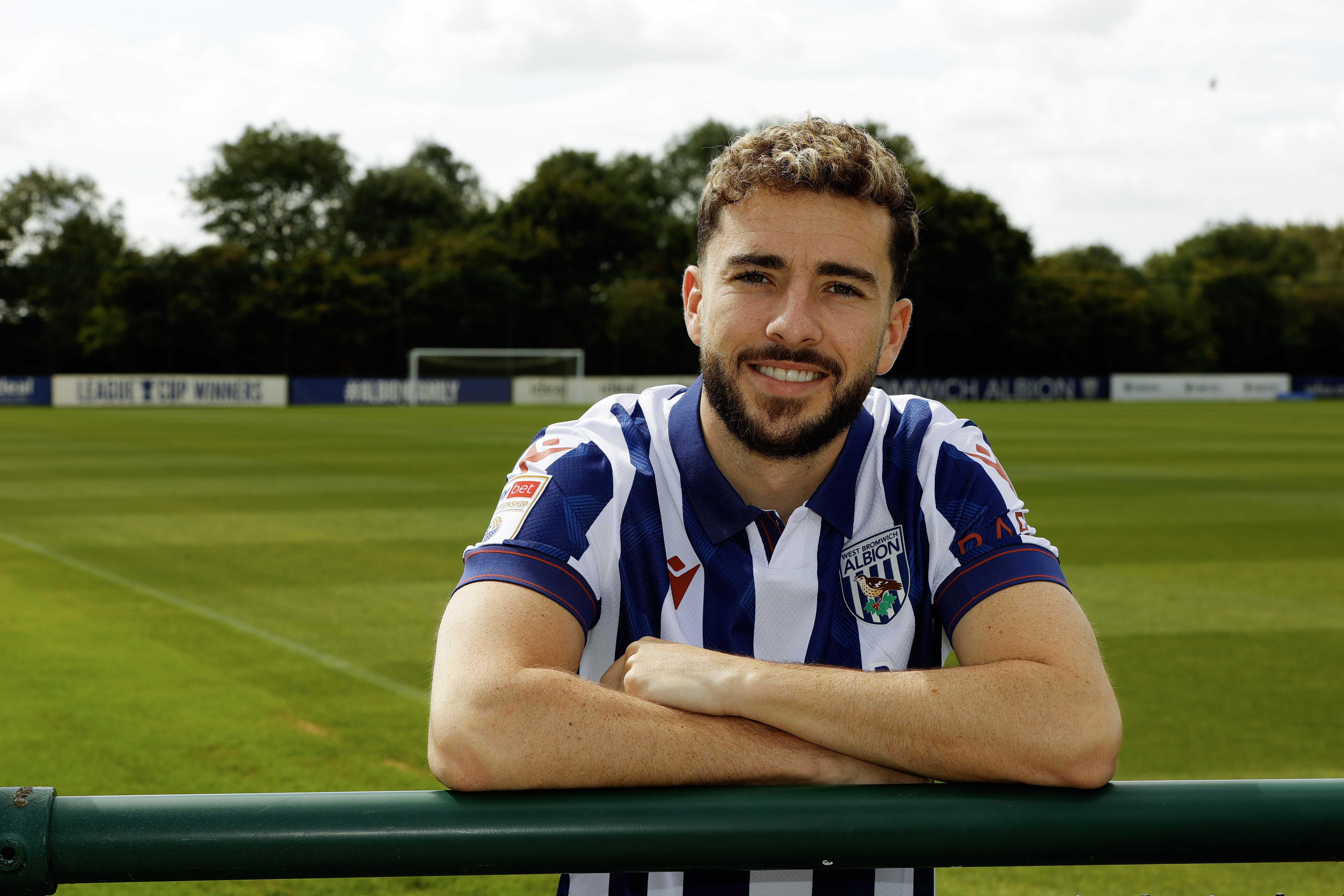 Mikey Johnston leaning over a barrier with his arms folded smiling at the camera while wearing a home shirt 