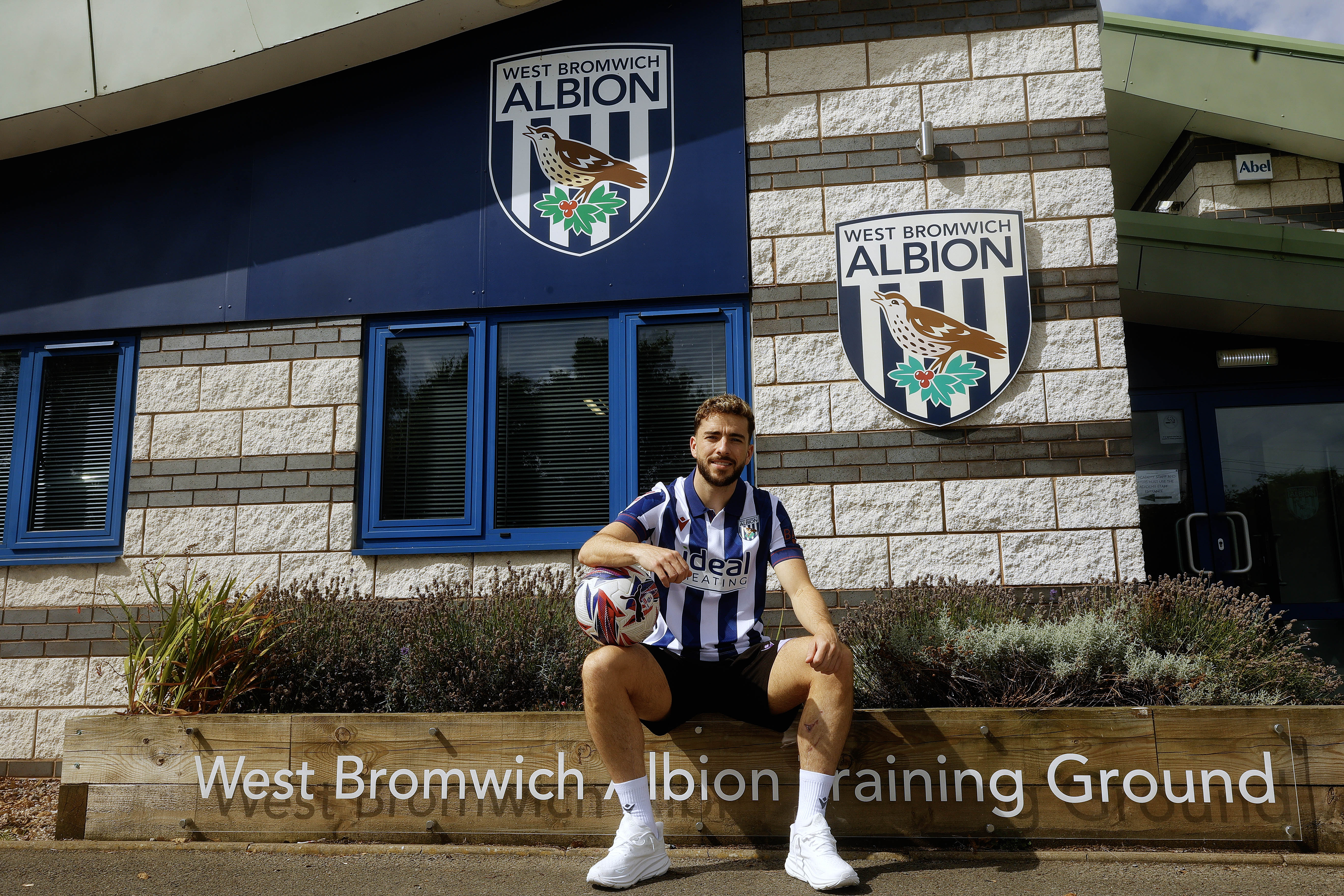 Mikey Johnston smiling at the camera while sat outside the training ground wearing a home shirt and wearing a ball