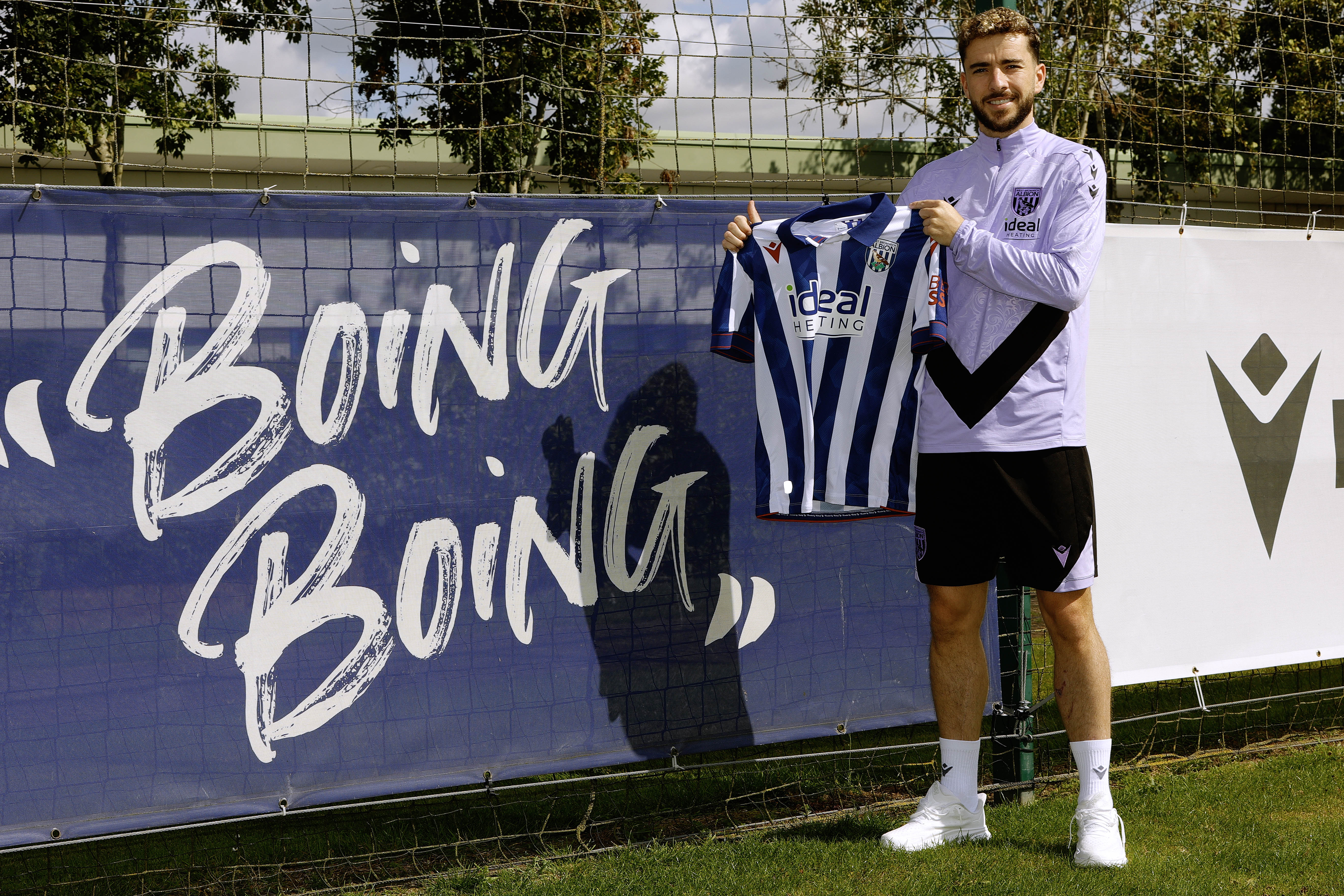 Mikey Johnston smiling at the camera holding up a home shirt stood next to a sign which says boing boing 