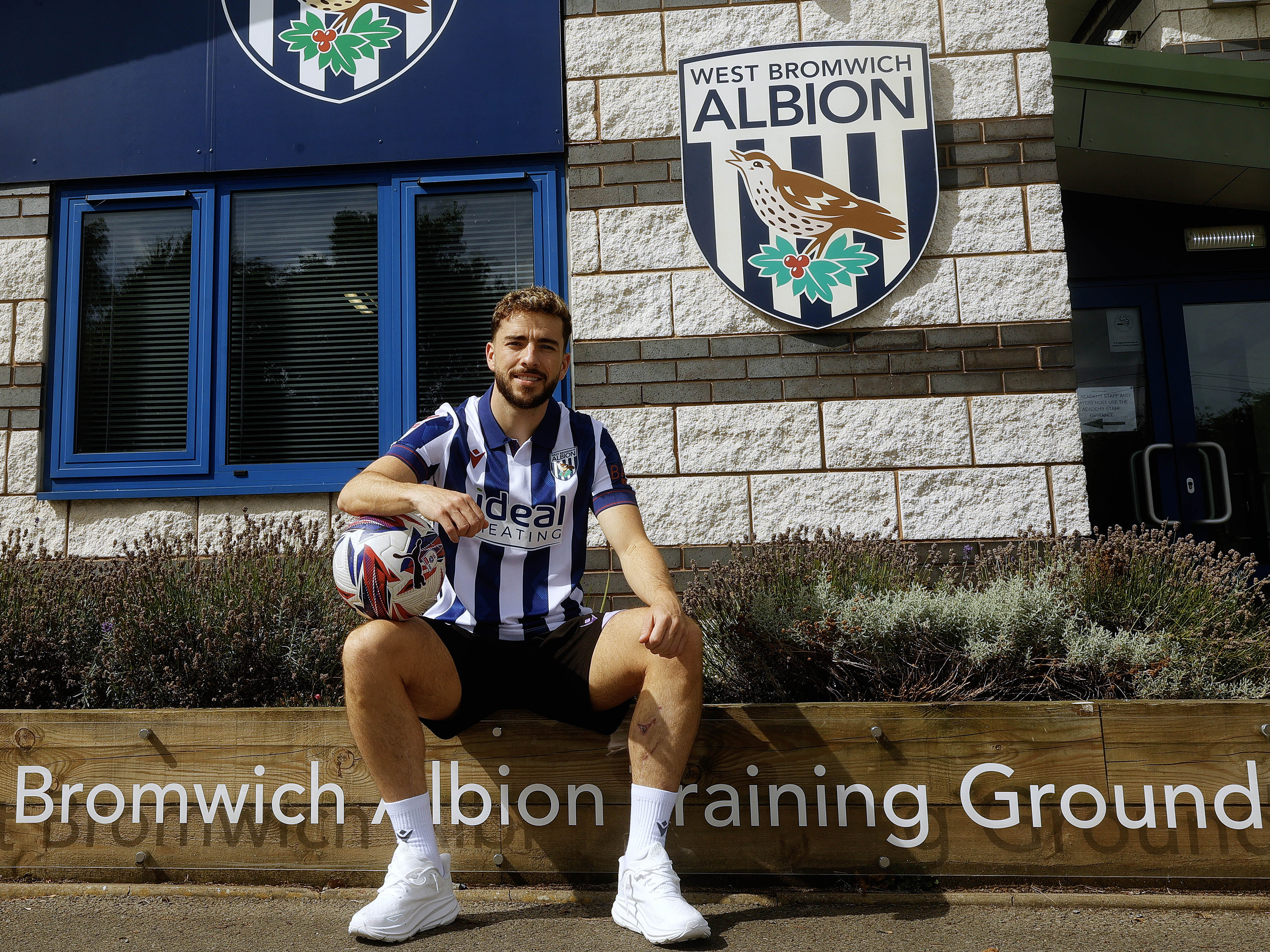 Mikey Johnston sat outside the training ground holding a ball and wearing the home shirt smiling at the camera 