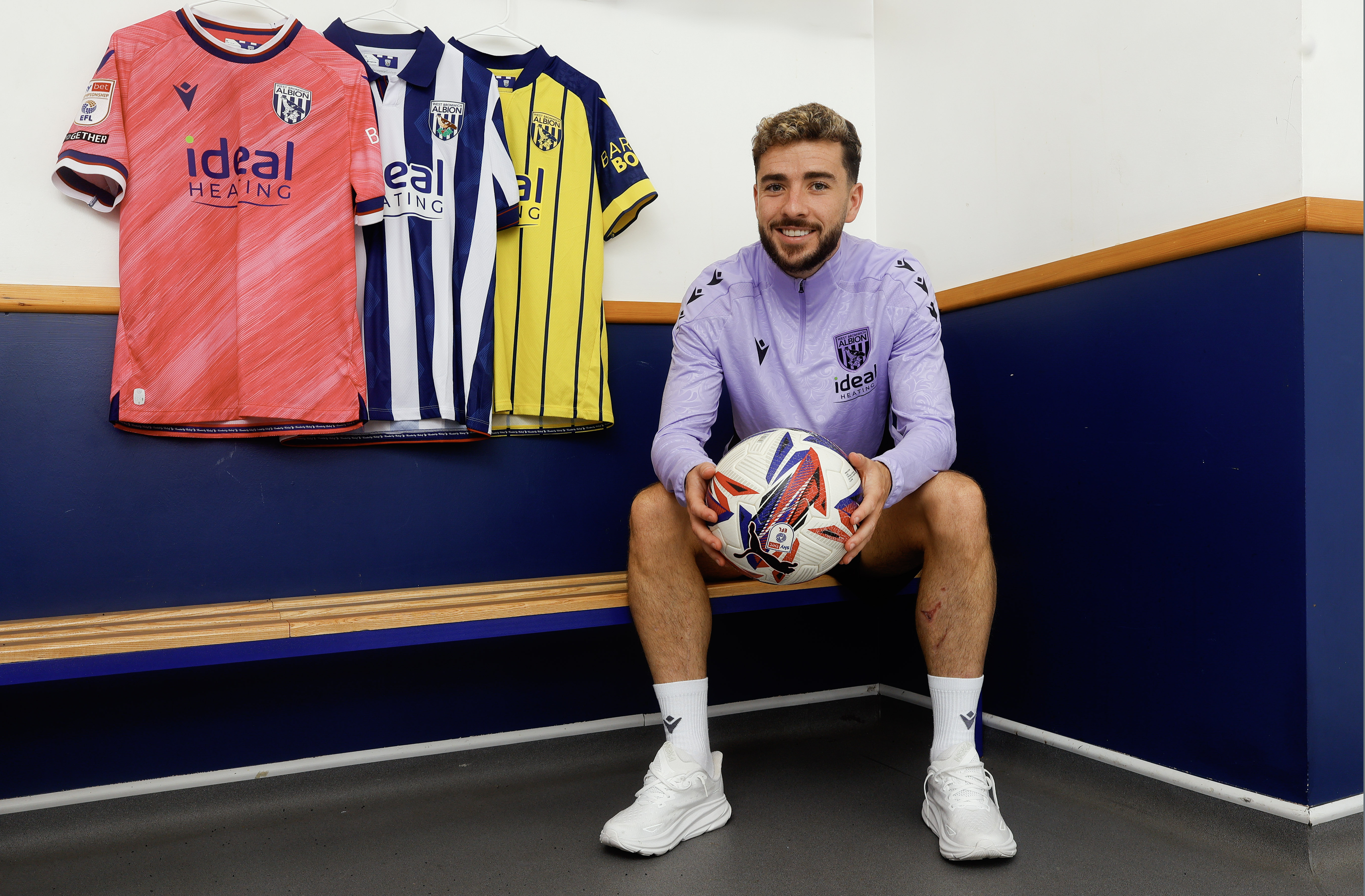 Mikey Johnston smiling at the camera while holding a ball while sat in a dressing room with all three 2024/25 shirts hanging up behind him