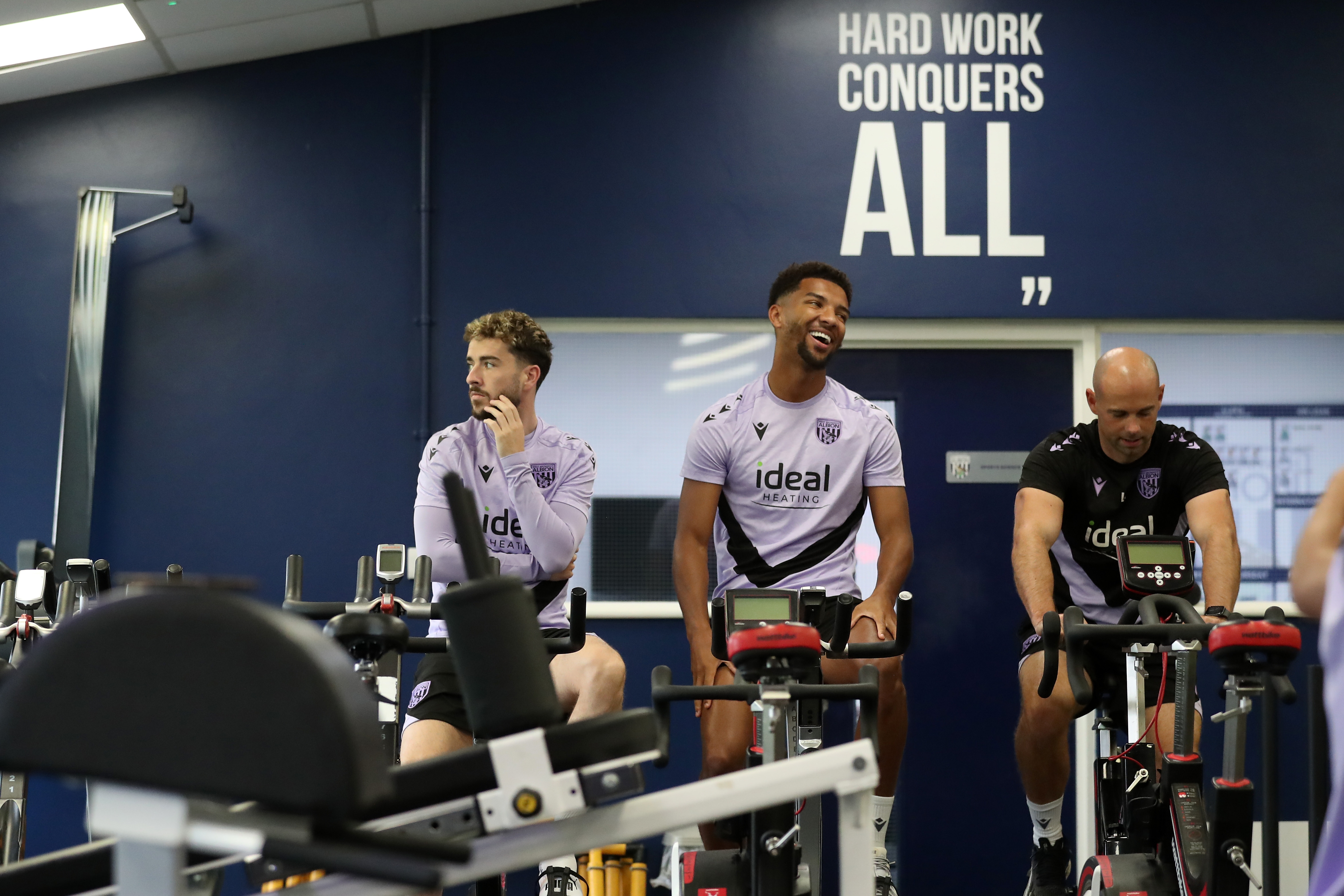 Mason Holgate on a bike in the gym sat next to Mikey Johnston and a member of staff with a smile on his face