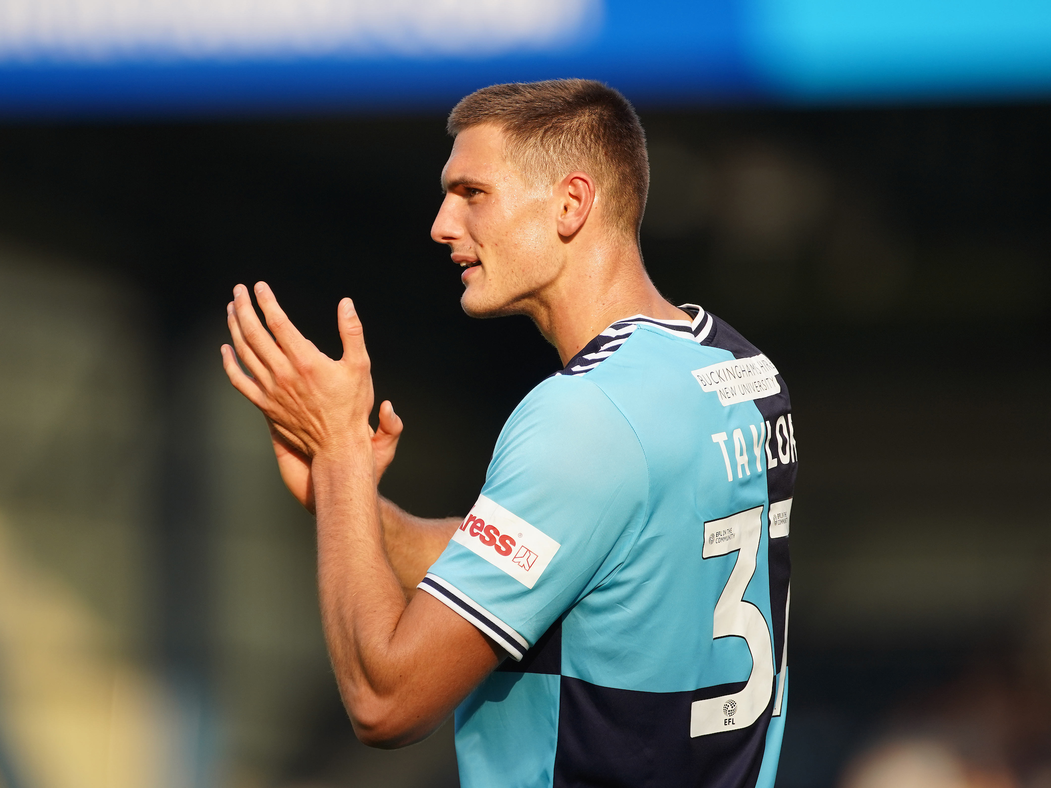 A photo of Caleb Taylor in the navy and sky blue home kit of loan club Wycombe, applauding supporters