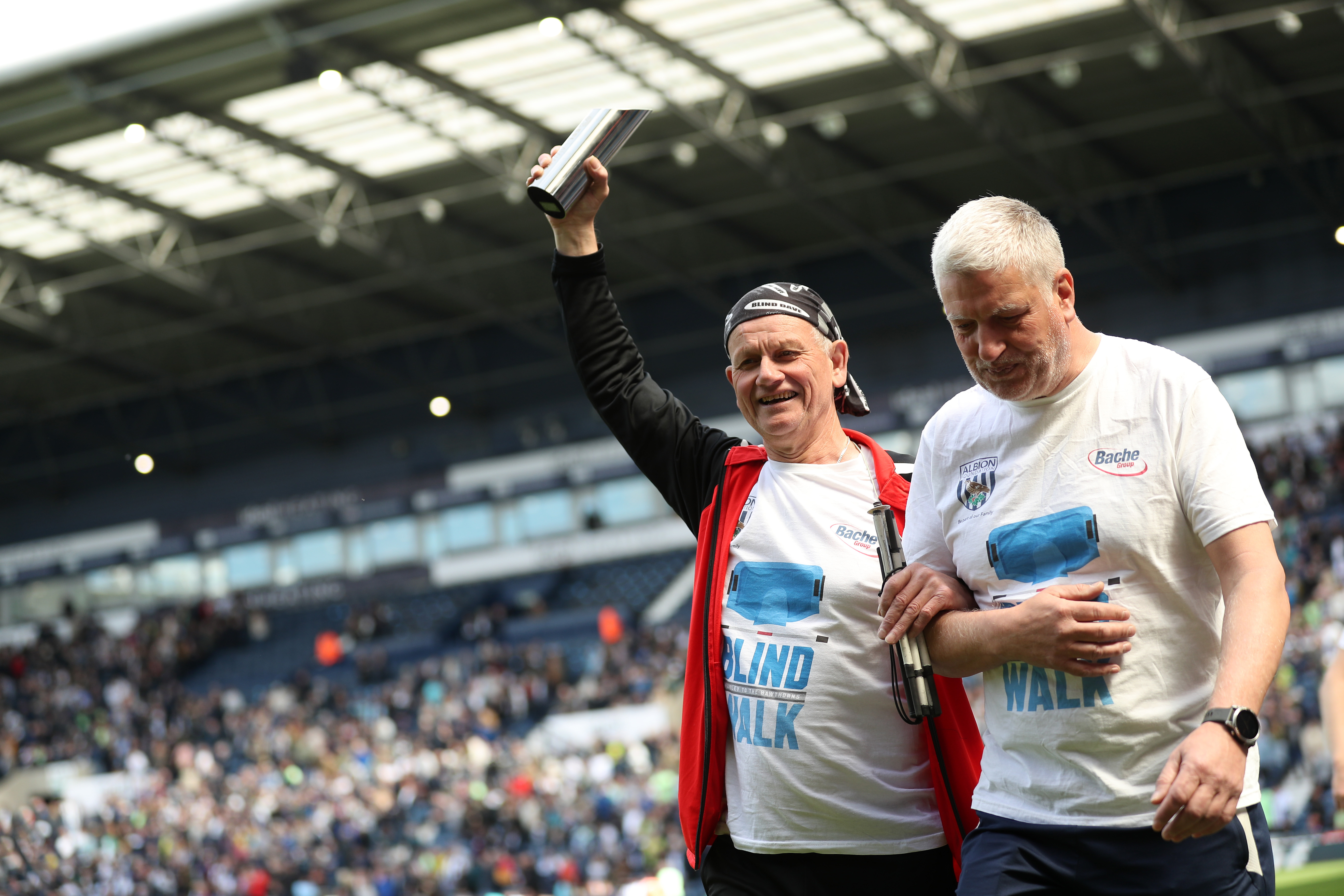 Tony Brown with his arm aloft walking around the side of the pitch at The Hawthorns with a member of Albion Foundation staff