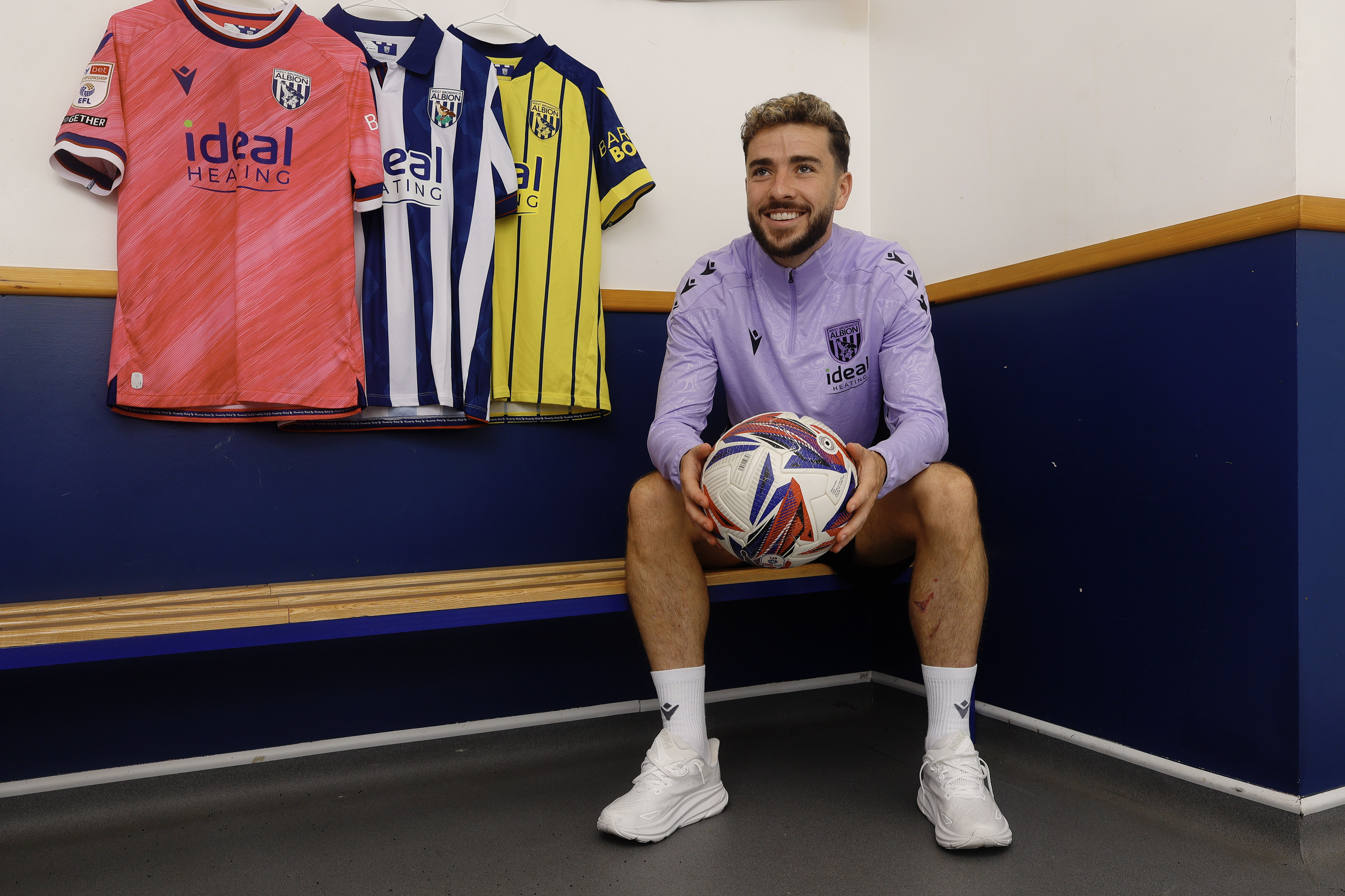 Mikey Johnston smiling while holding a ball in a changing room with three shirts hanging above his head 