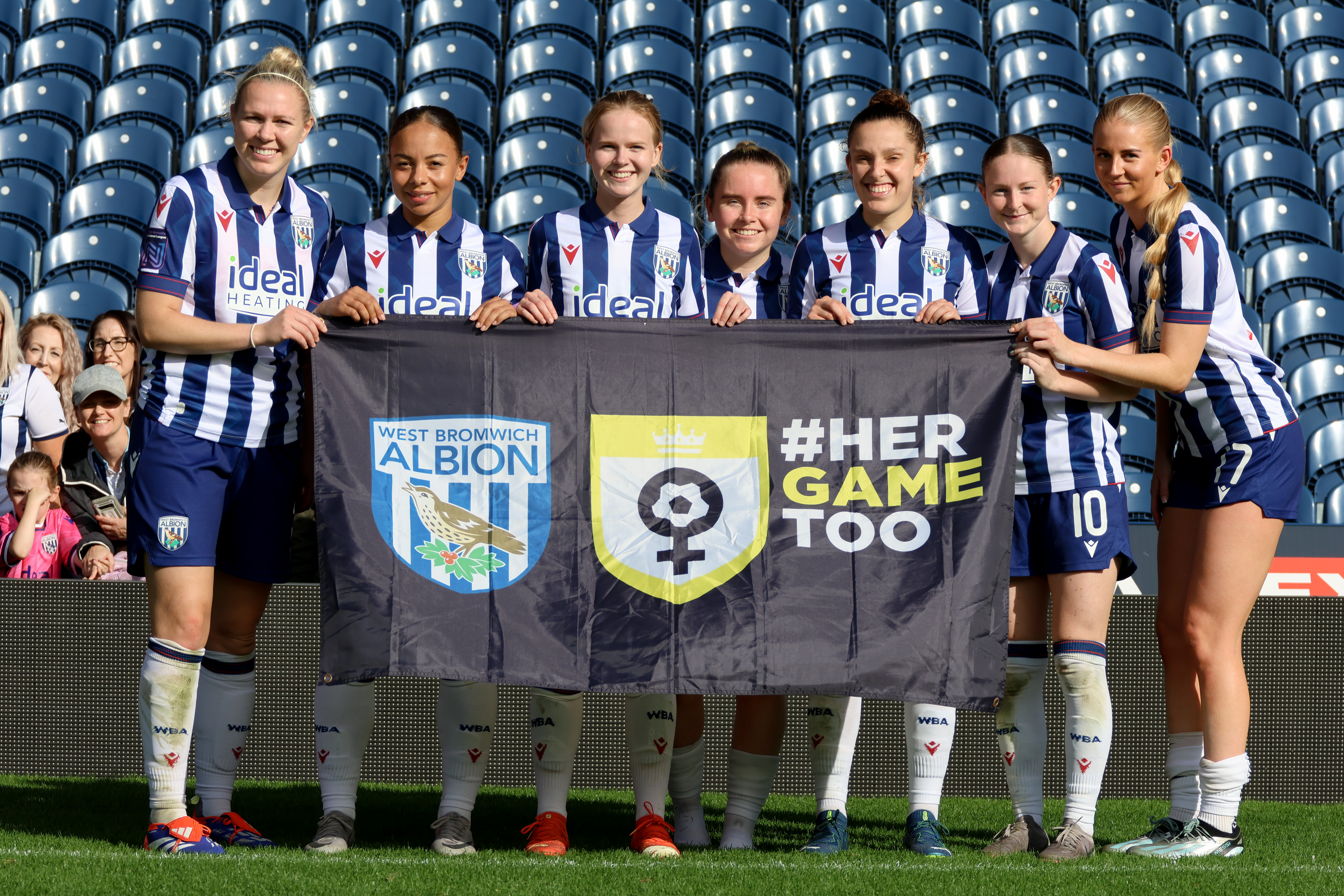 Several Albion Women Player hold up a Her Game Too flag at The Hawthorns while in home kit 