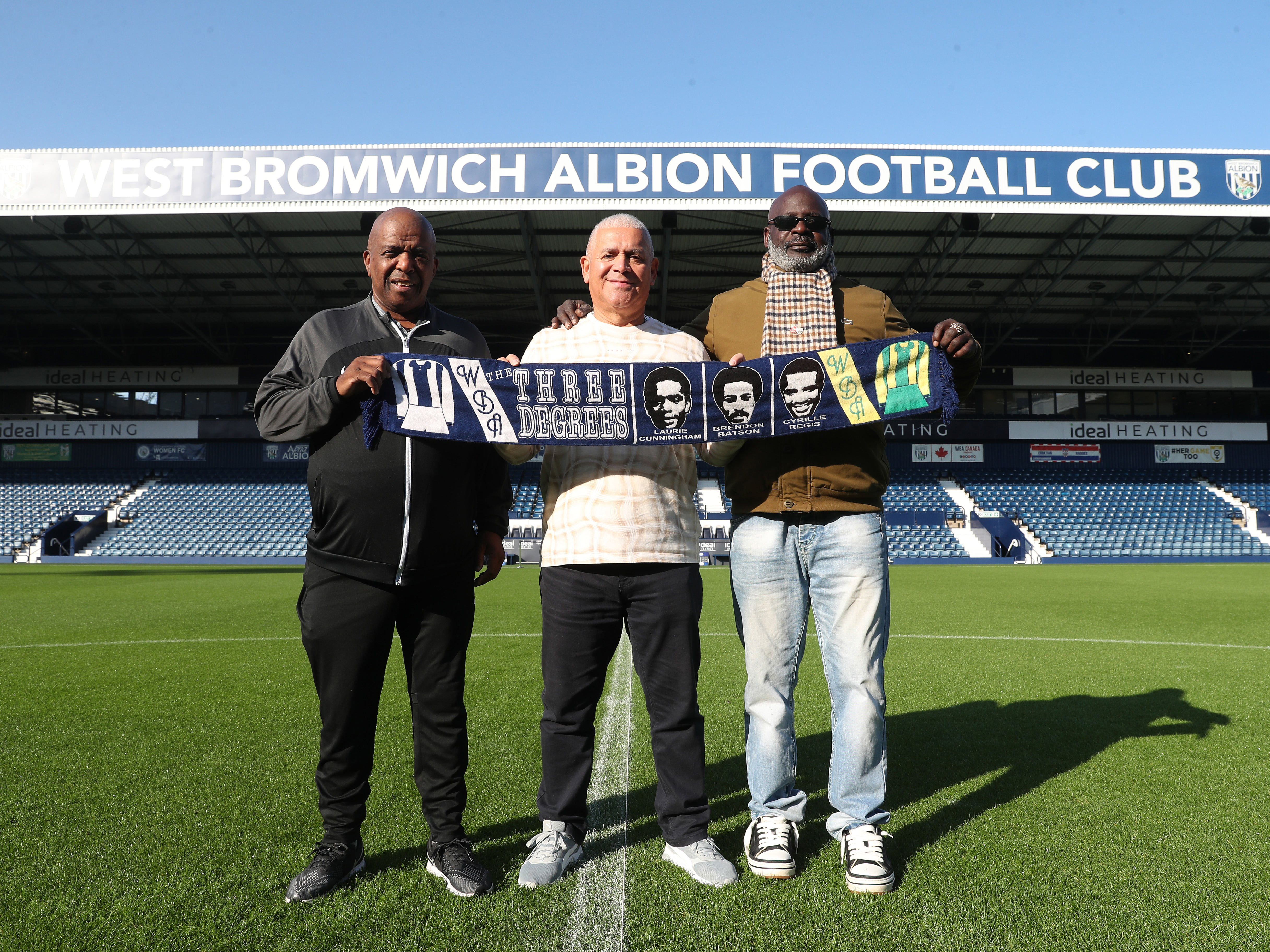 Three Albion supporters hold a WBA scarf while stood on the centre circle at The Hawthorns in front of the Halfords Lane Stand 